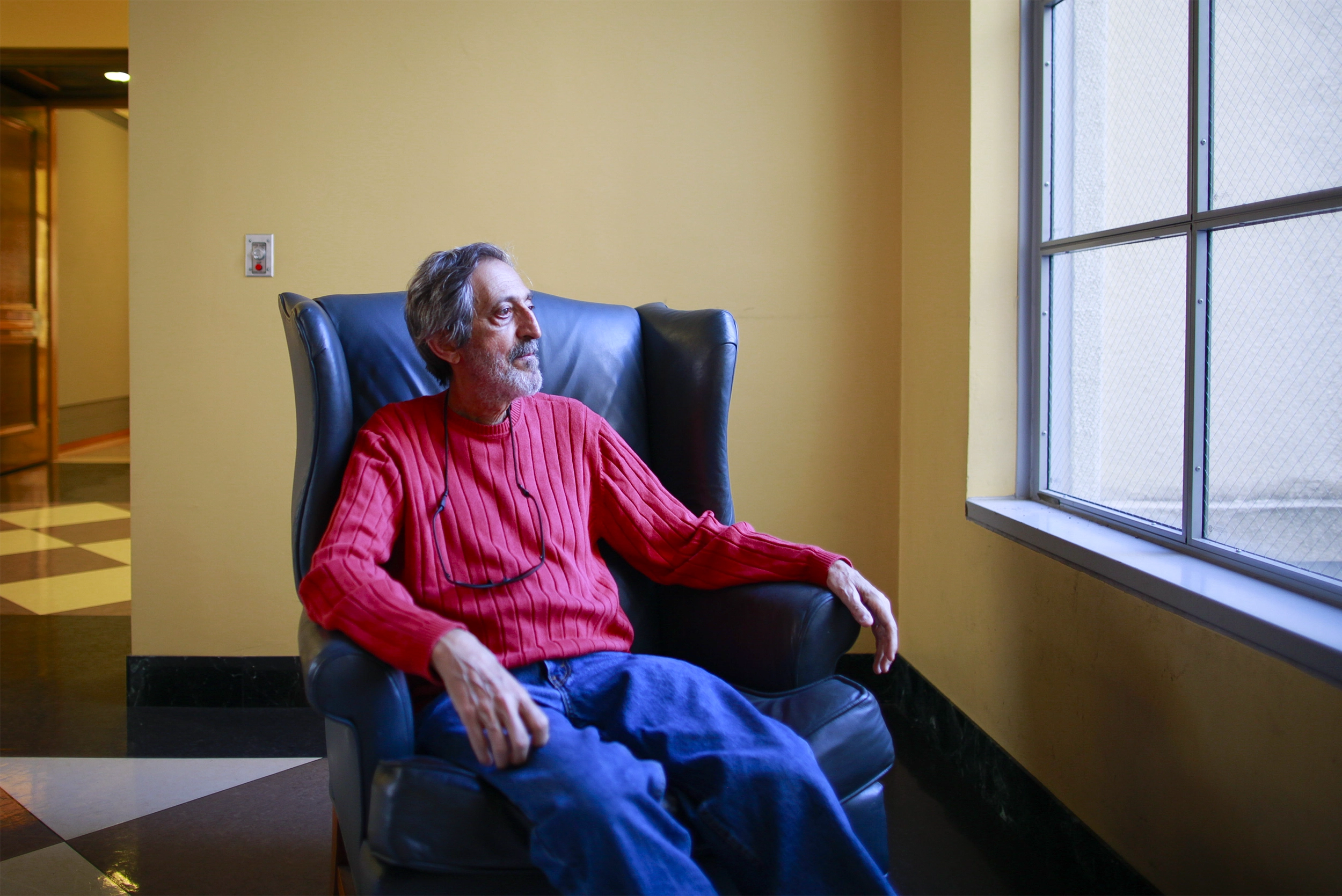 Avi Wigderson in a red sweater and blue jeans sits in a black chair and looks out a window.