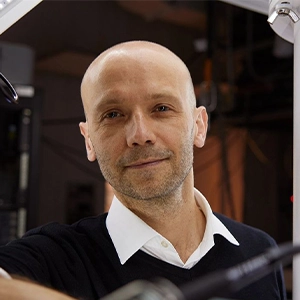 Close-up portrait of a man leaning against some equipment in a lab and smiling at the camera.