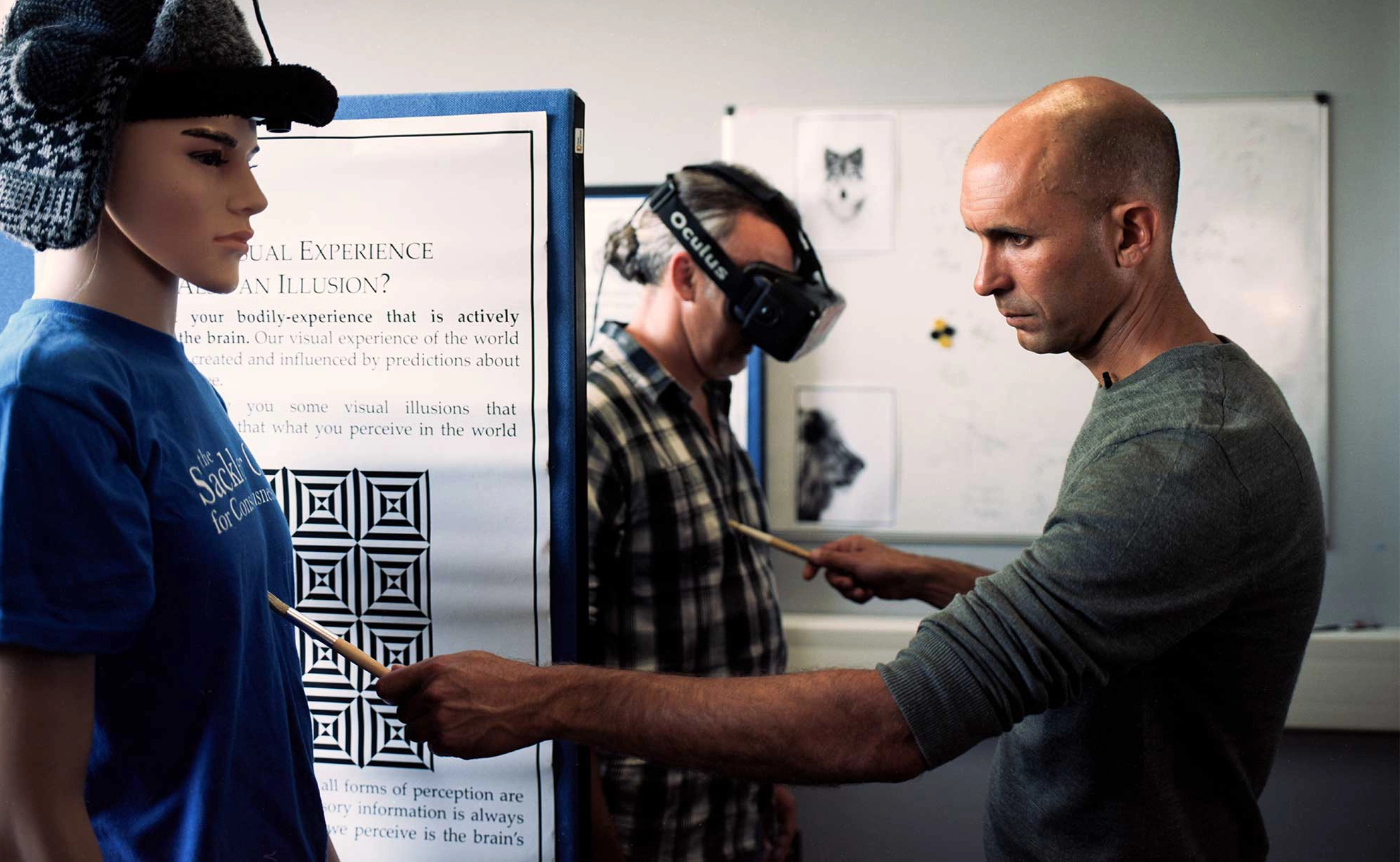 A man in profile strokes a paintbrush on the chests of a man in a VR head and a manequin.