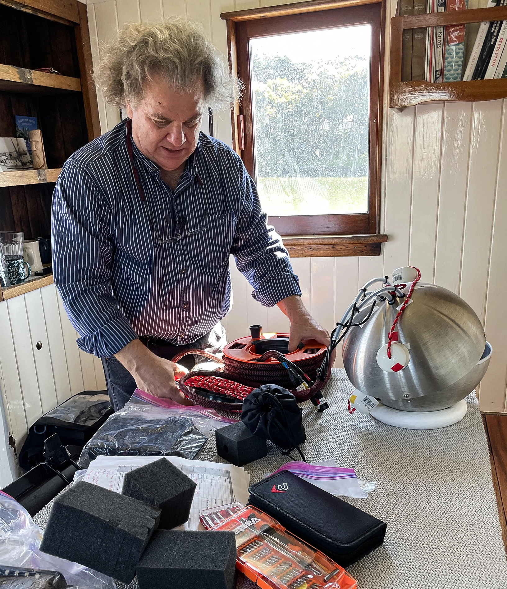 A man with gray hair stands next to an array of gadgets on a table, including a stainless-steel sphere with wires coming out of it.