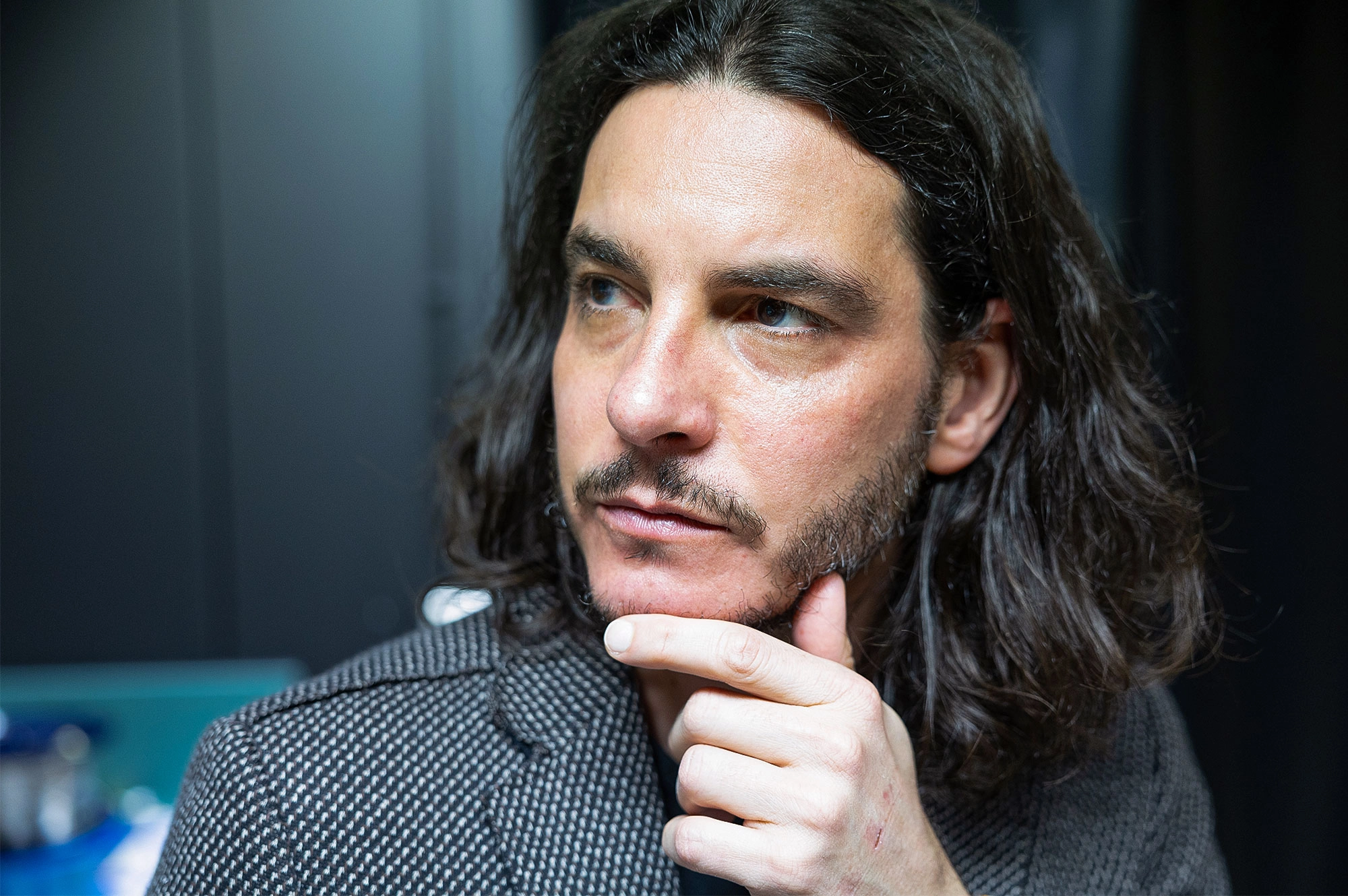 Close-up of a man’s face. He has long, dark, wavy hair and a close-clipped beard and mustache.