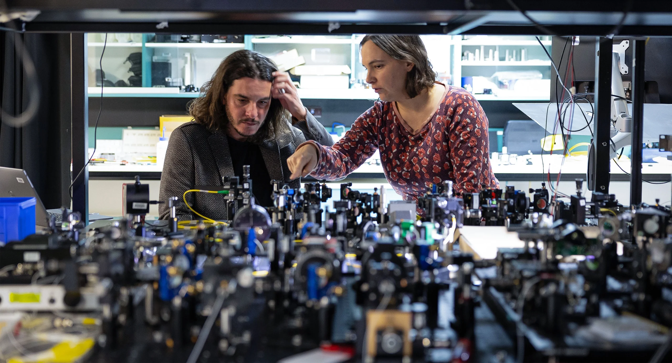 A man and woman converse while peering down at an experimental setup on an optical table.