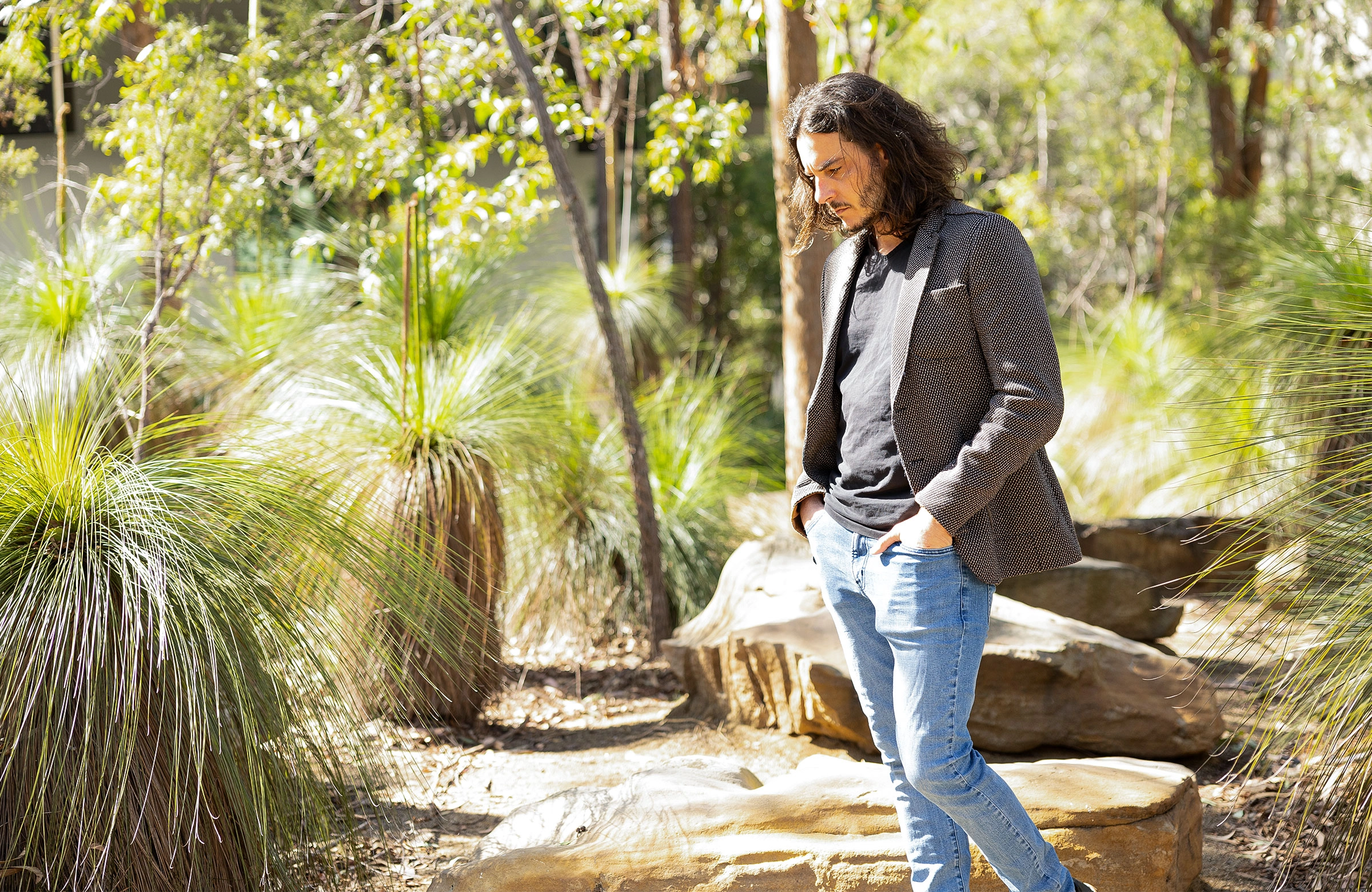 A man in a blazer with long dark hair gazes at the ground with his hands in his jeans pockets, amid trees.