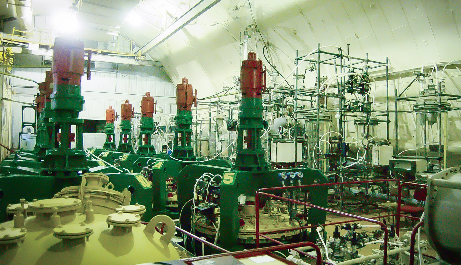 A yellow-lit laboratory crammed with equipment, including a set of 10 chemical reactors with red caps.