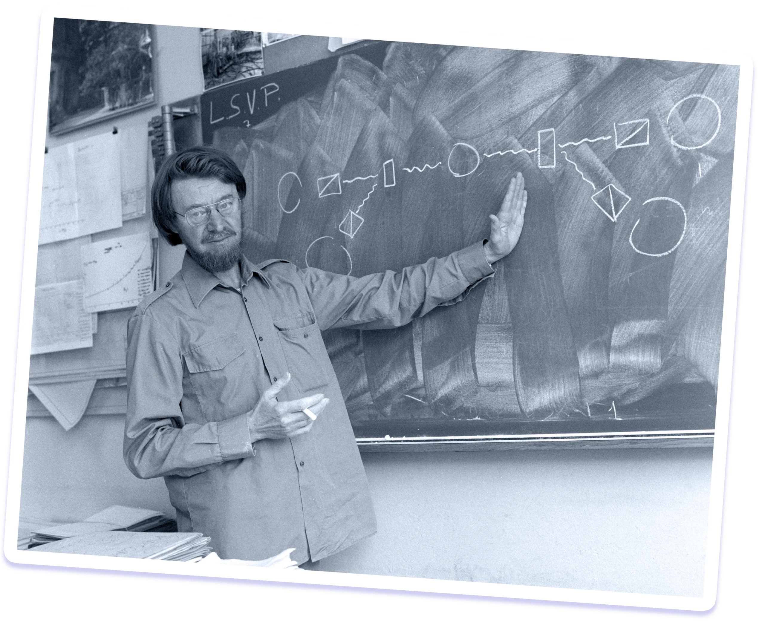 Black-and-white photo of a man with a mustache and beard gesturing to a drawing on a blackboard that shows the design of an experiment.