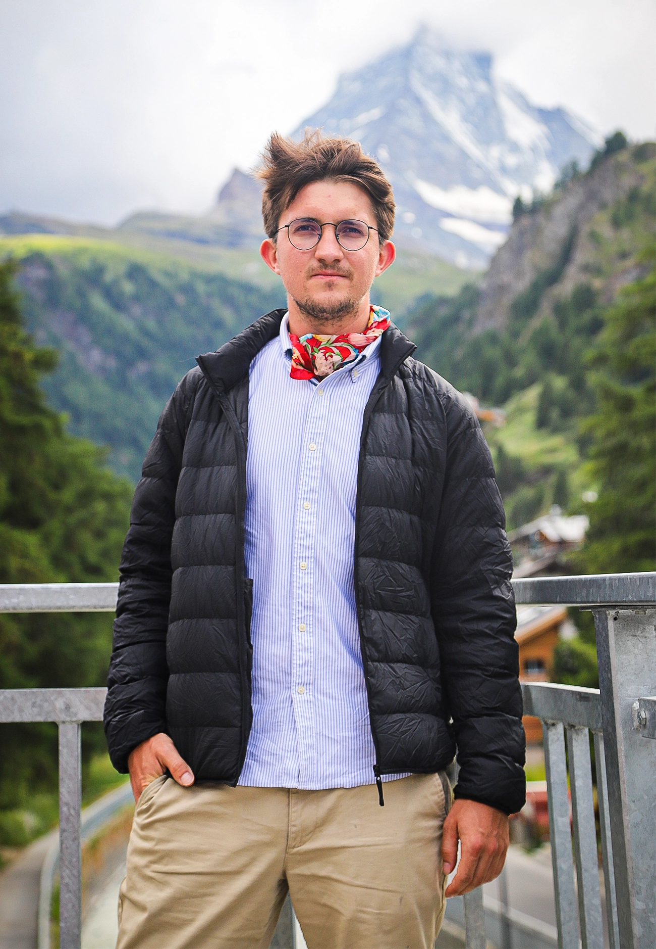 A young man wearing a red scarf stands in front of a mountain.