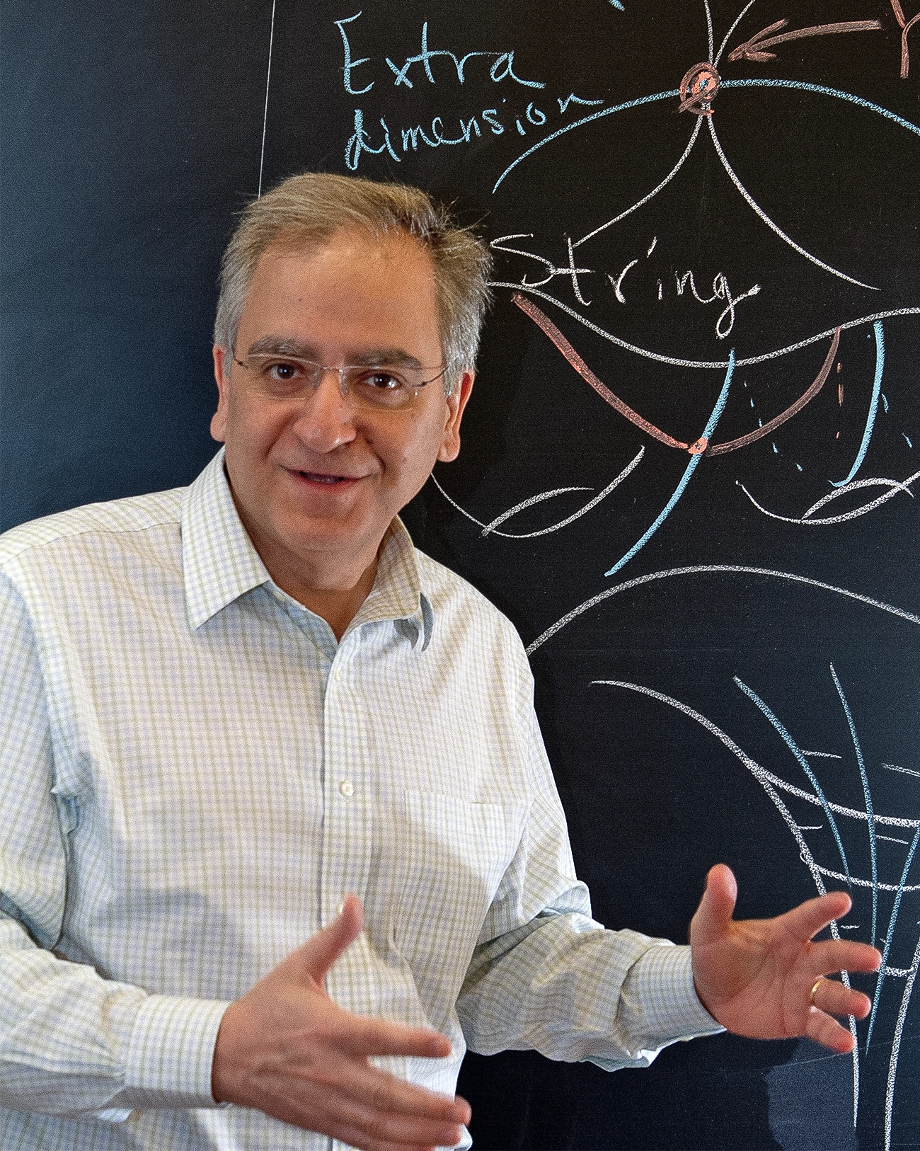 A man in a white shirt gestures as if he is explaining something while standing in front of a blackboard.