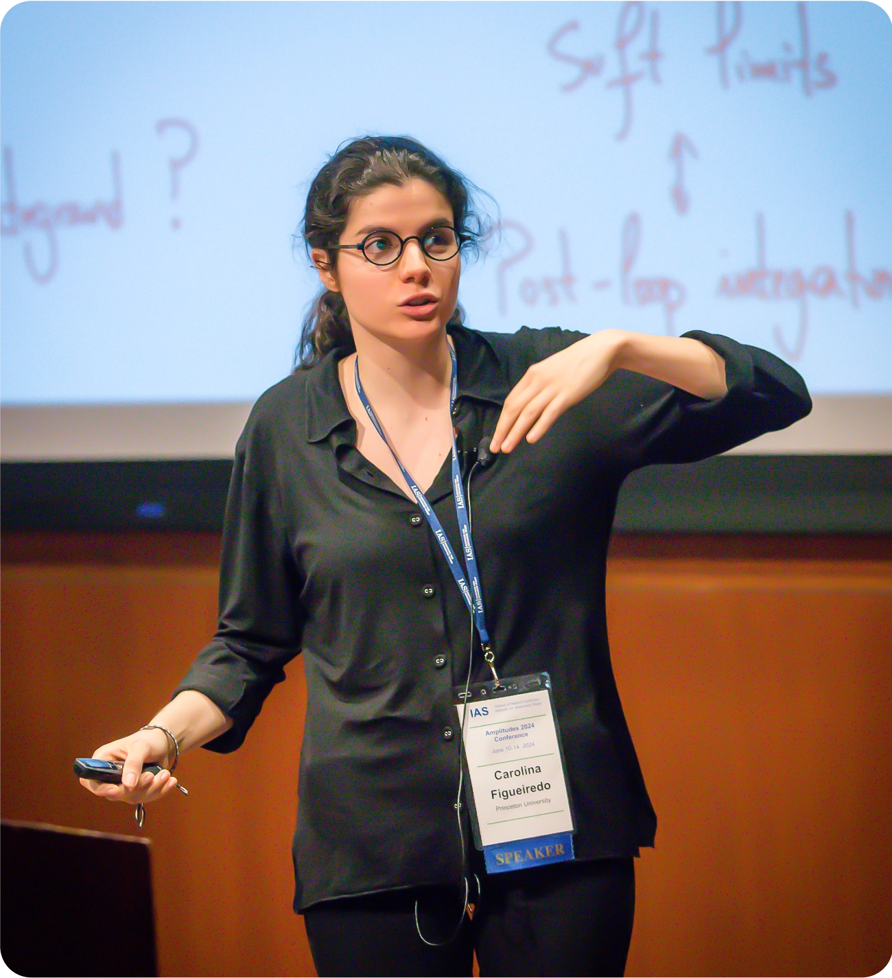 A woman speaks on a stage. She stands in front of a presentation slide and carries a clicker.