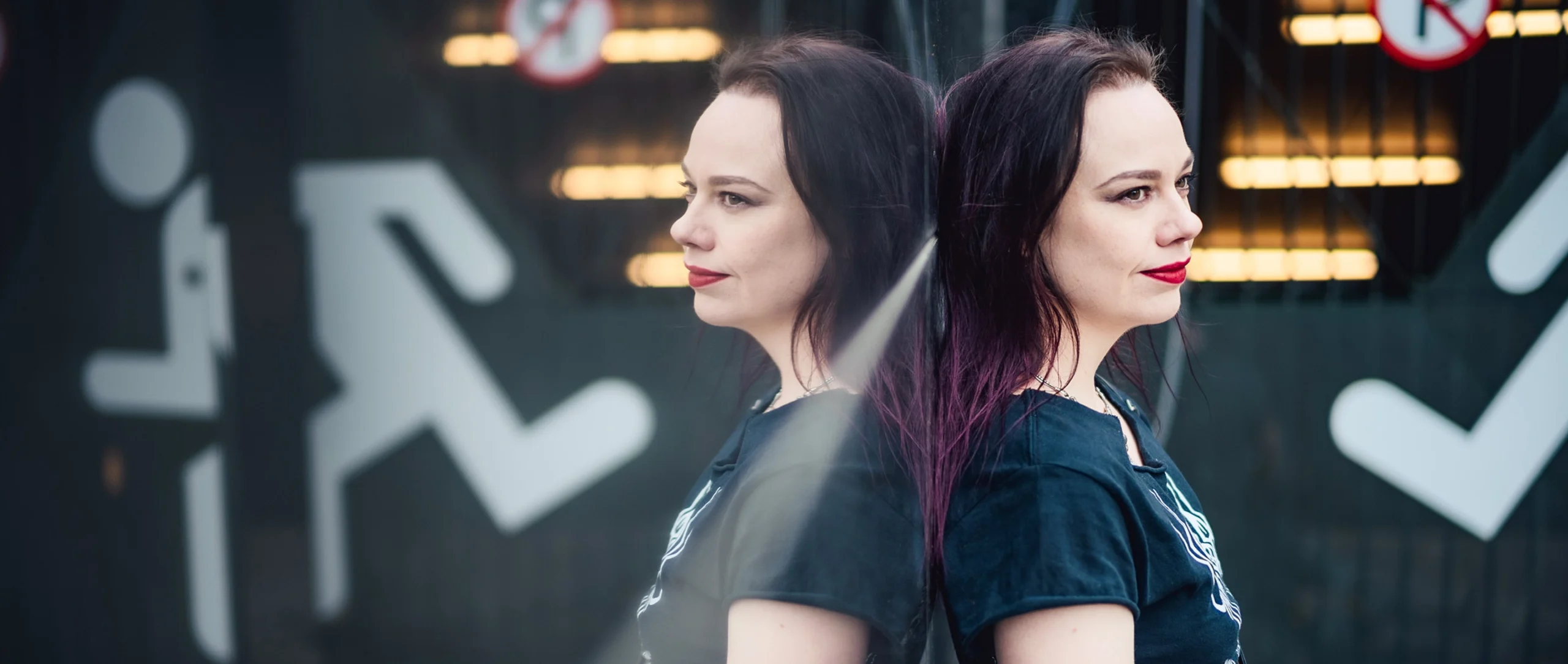 A woman with black hair in a black T-shirt leaning against a mirror and seen in reflection.