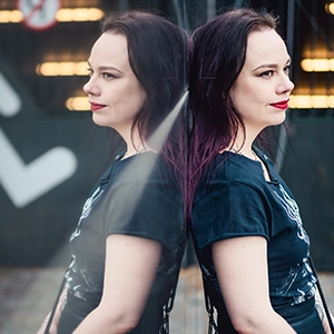 A woman with black hair in a black T-shirt leaning against a mirror and seen in reflection.