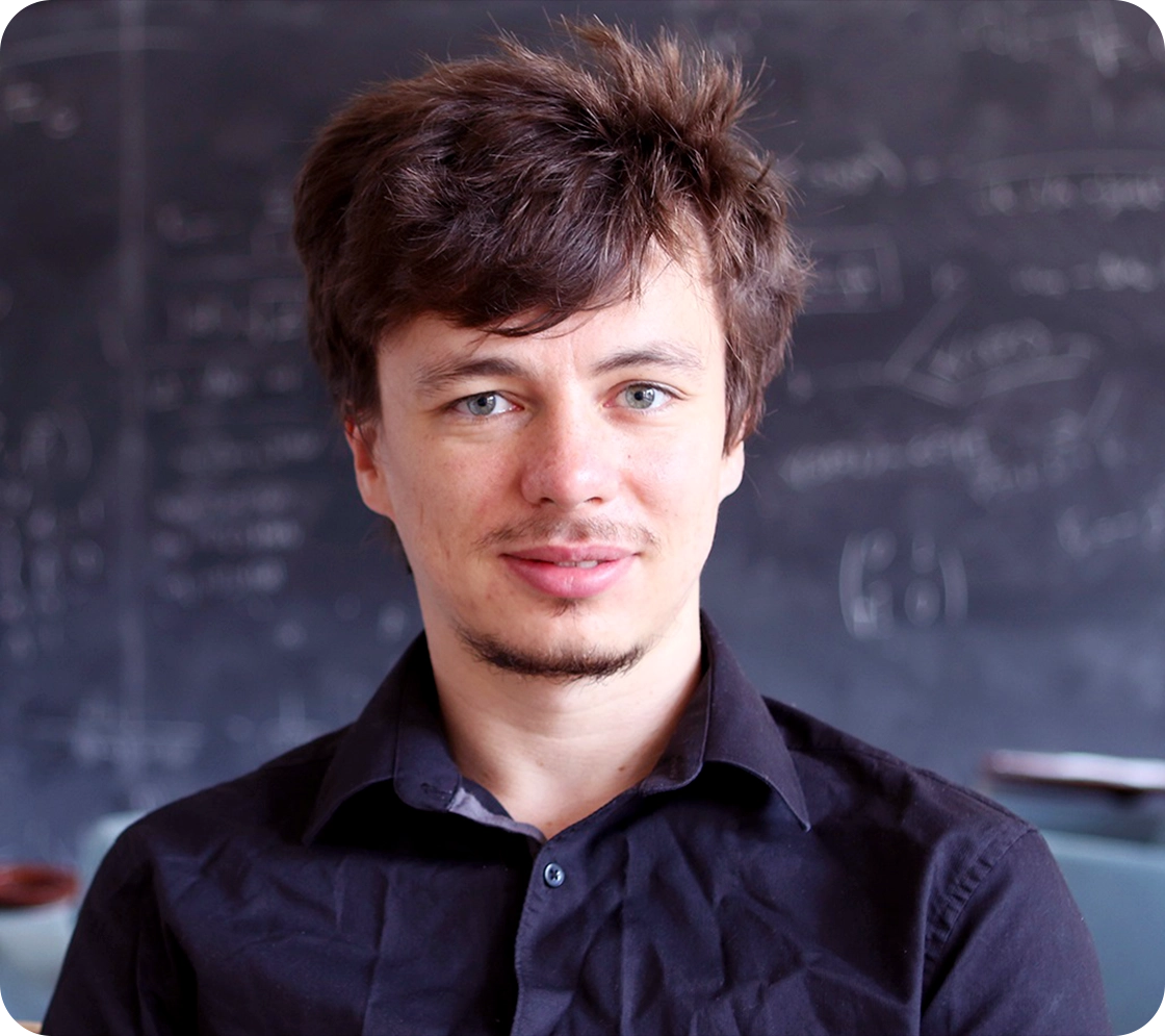 A man with disheveled hair stands in front of a chalkboard.