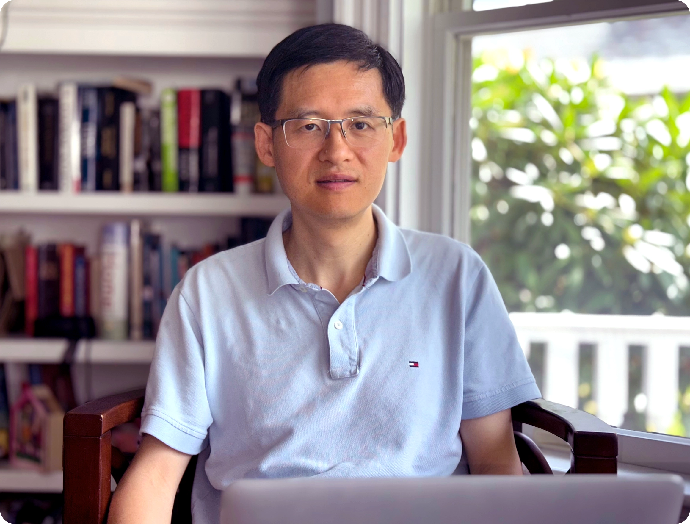 A man sits in a sunny room in front of a bookshelf.