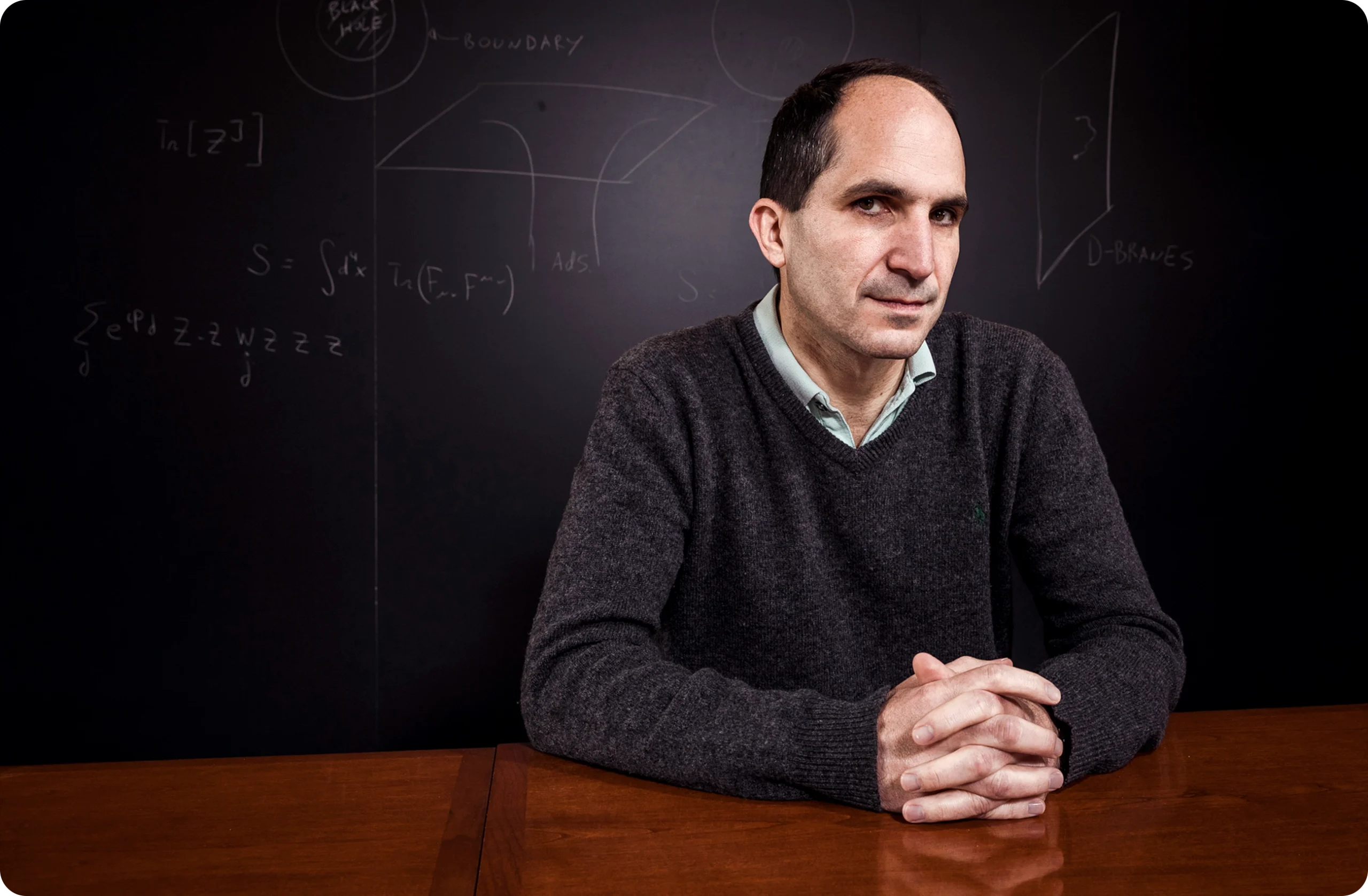 A man sits at a wooden table with a blackboard in the background.