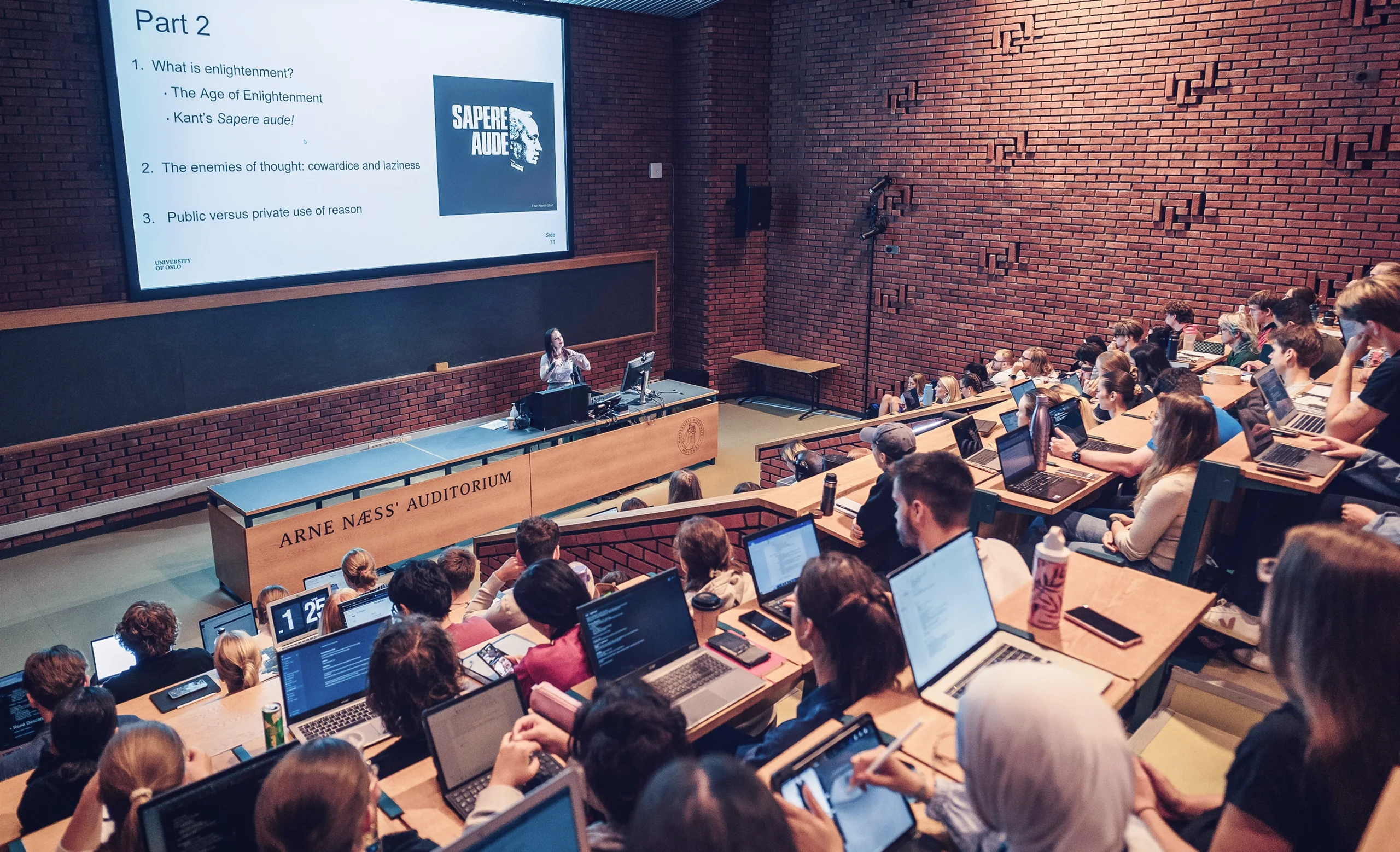 A woman stands at a podium in a large lecture hall full of students. Her slide asks, “What is enlightenment?”