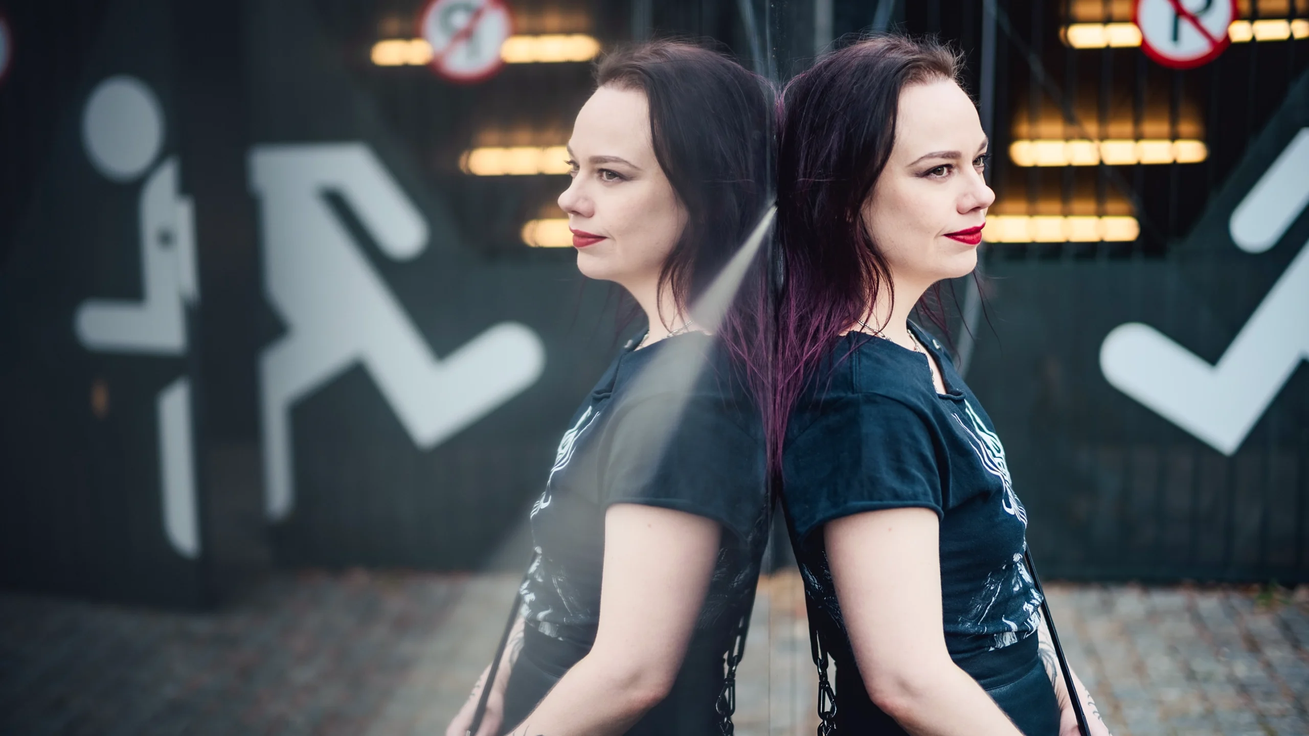 A woman with black hair in a black T-shirt leaning against a mirror and seen in reflection.