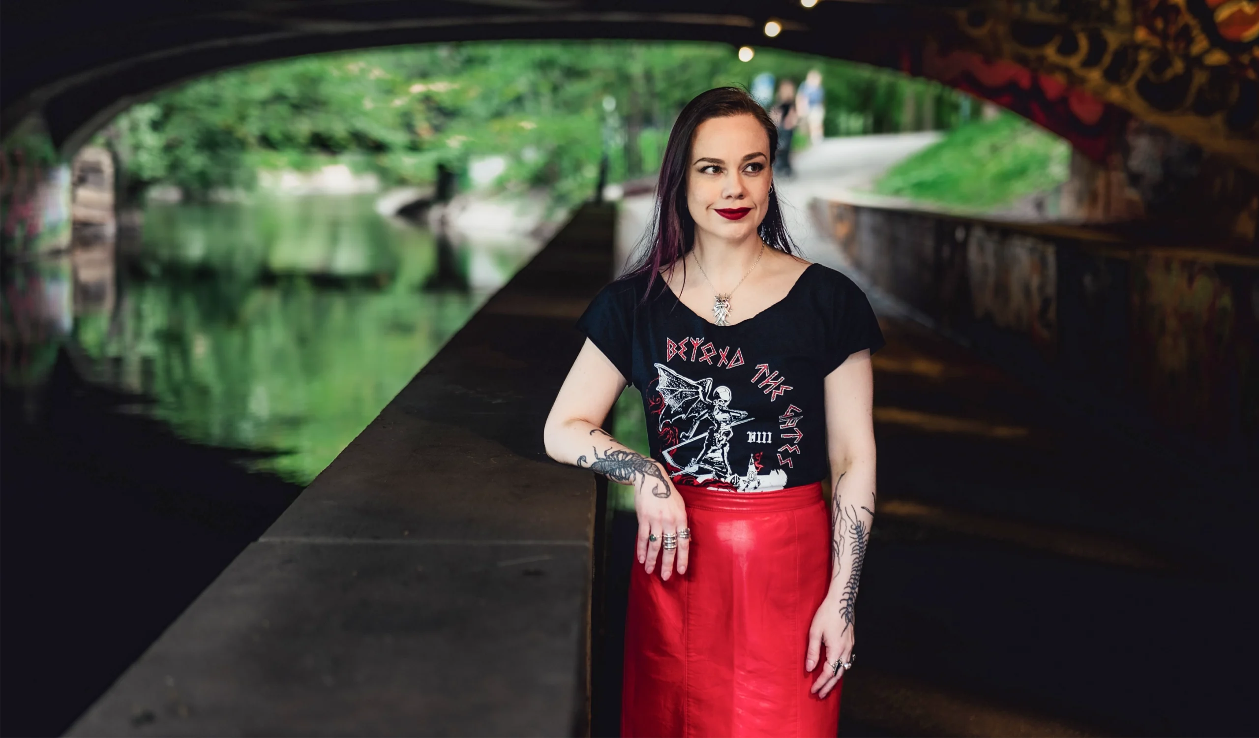 A woman with black hair and tattoos stands under a dark bridge with a verdant, tree-filled scene behind her.