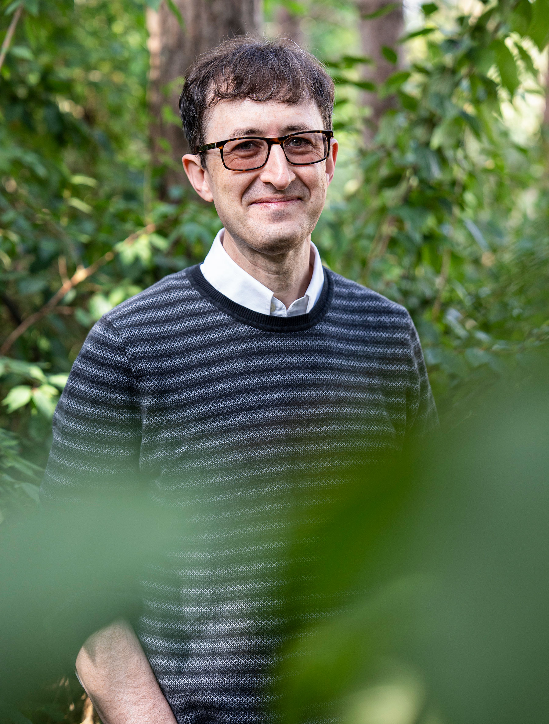 A man stands and smiles at the camera while standing in front of some trees.