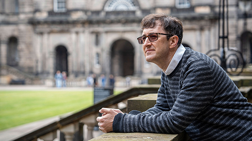 A man in variable-tint glasses leans on a stone balustrade overlooking a lawn at the University of Edinburgh.