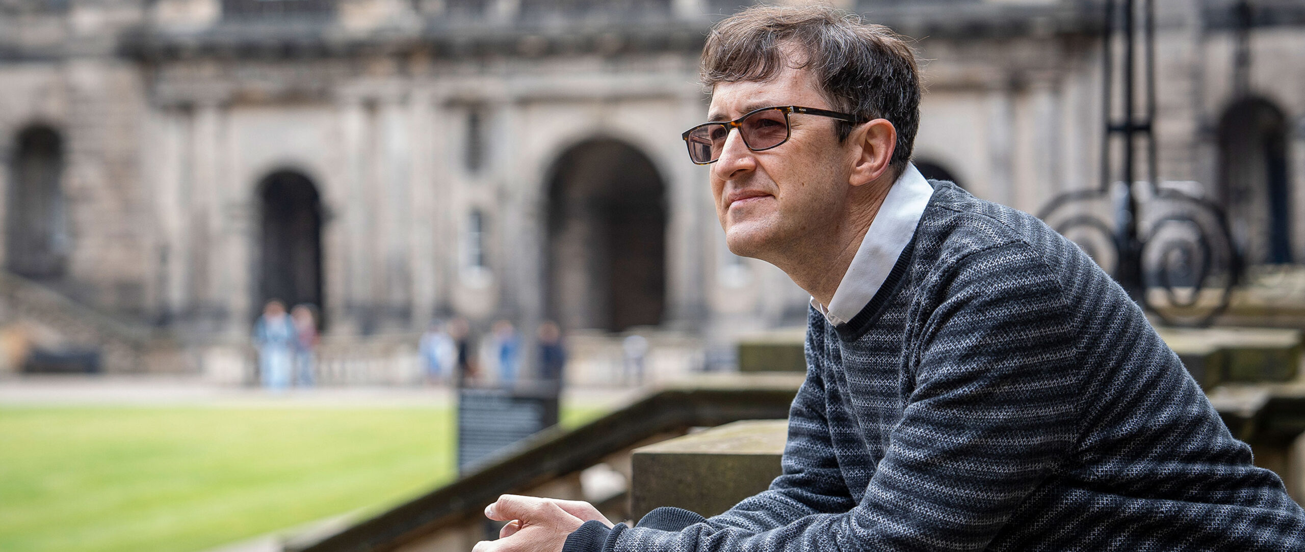 A man in variable-tint glasses leans on a stone balustrade overlooking a lawn at the University of Edinburgh.