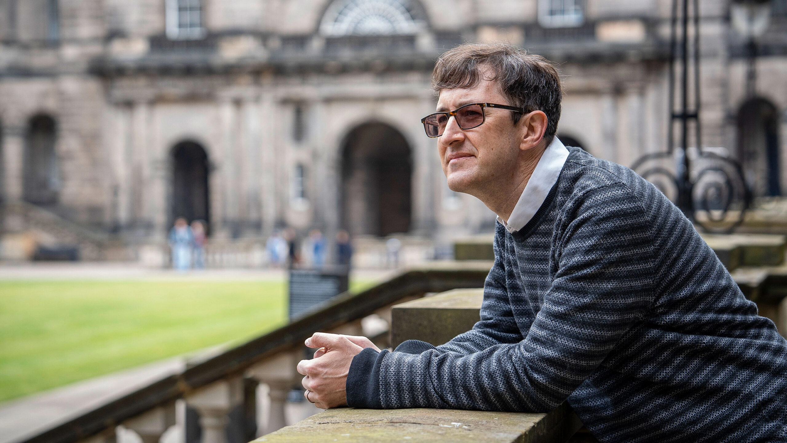 A man in variable-tint glasses leans on a stone balustrade overlooking a lawn at the University of Edinburgh.