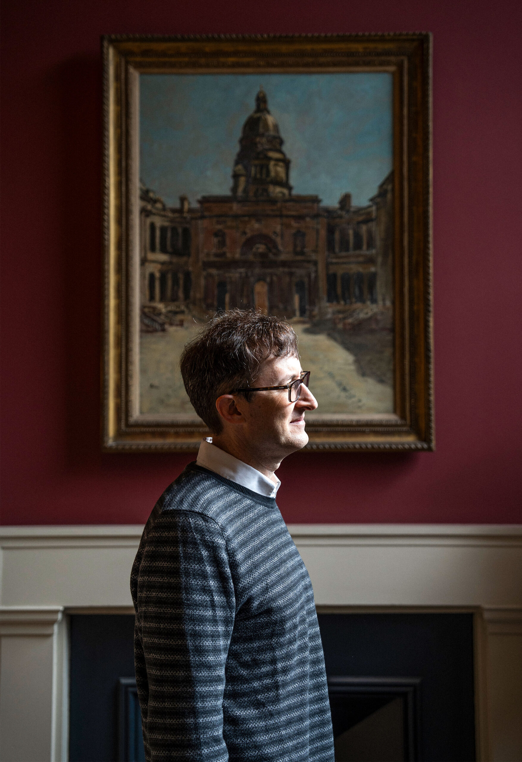 A man is seen in profile in front of an oil painting of a building.