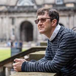 A man in variable-tint glasses leans on a stone balustrade overlooking a lawn at the University of Edinburgh.