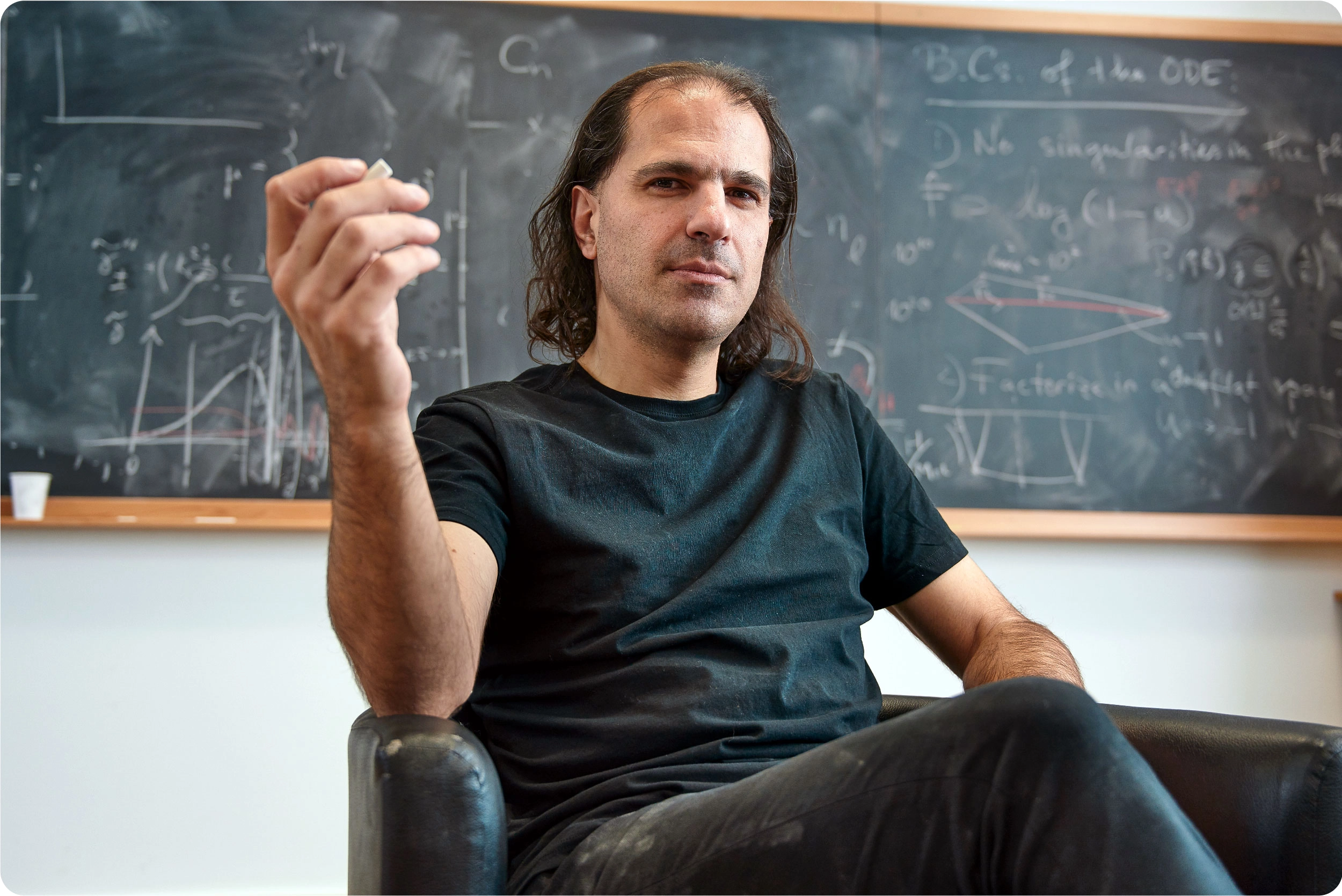 A man sits in front of a blackboard, holding a piece of chalk.