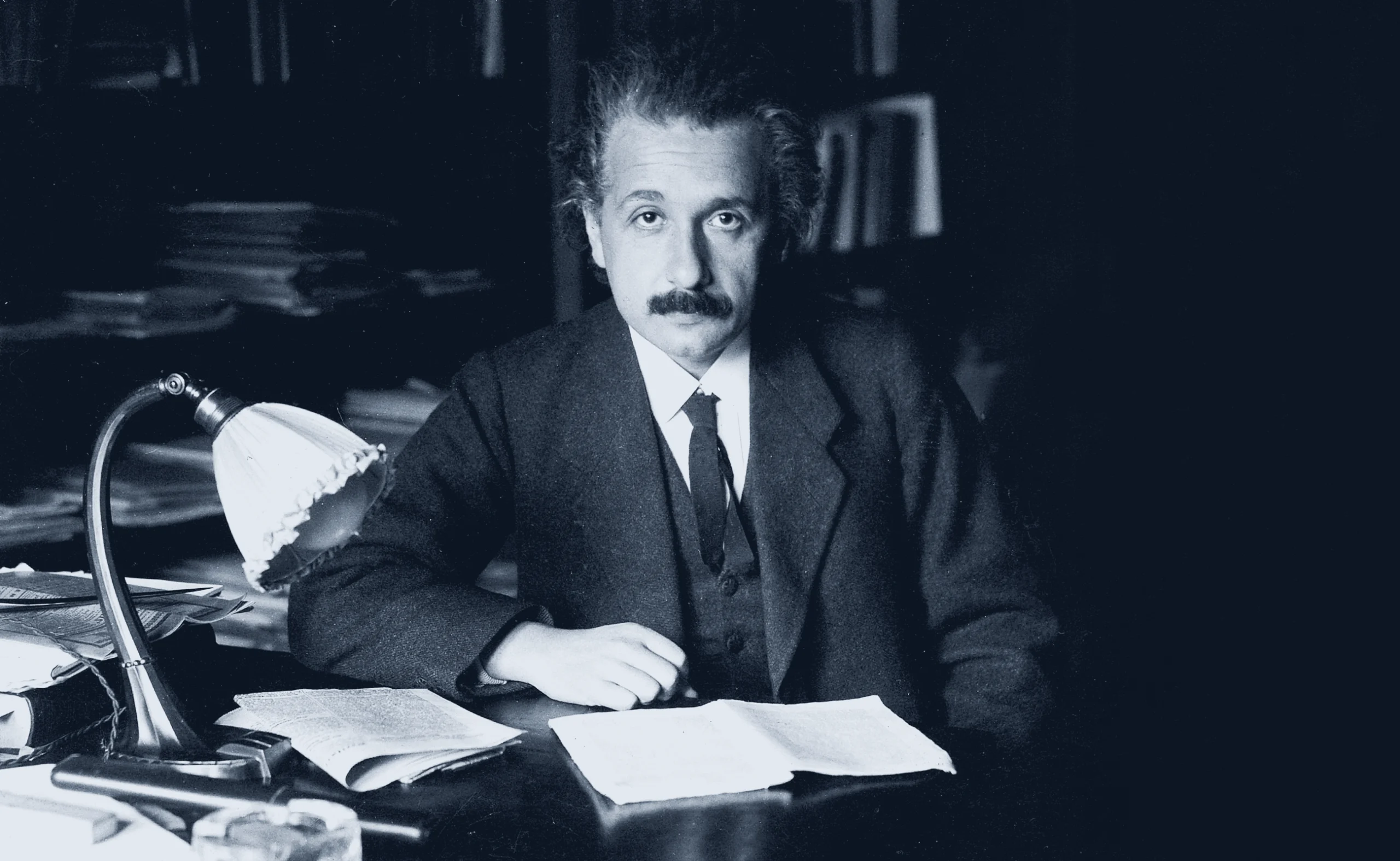 Black-and-white blue tinted photo of Albert Einstein sitting at his desk looking serious in a three-piece suit.