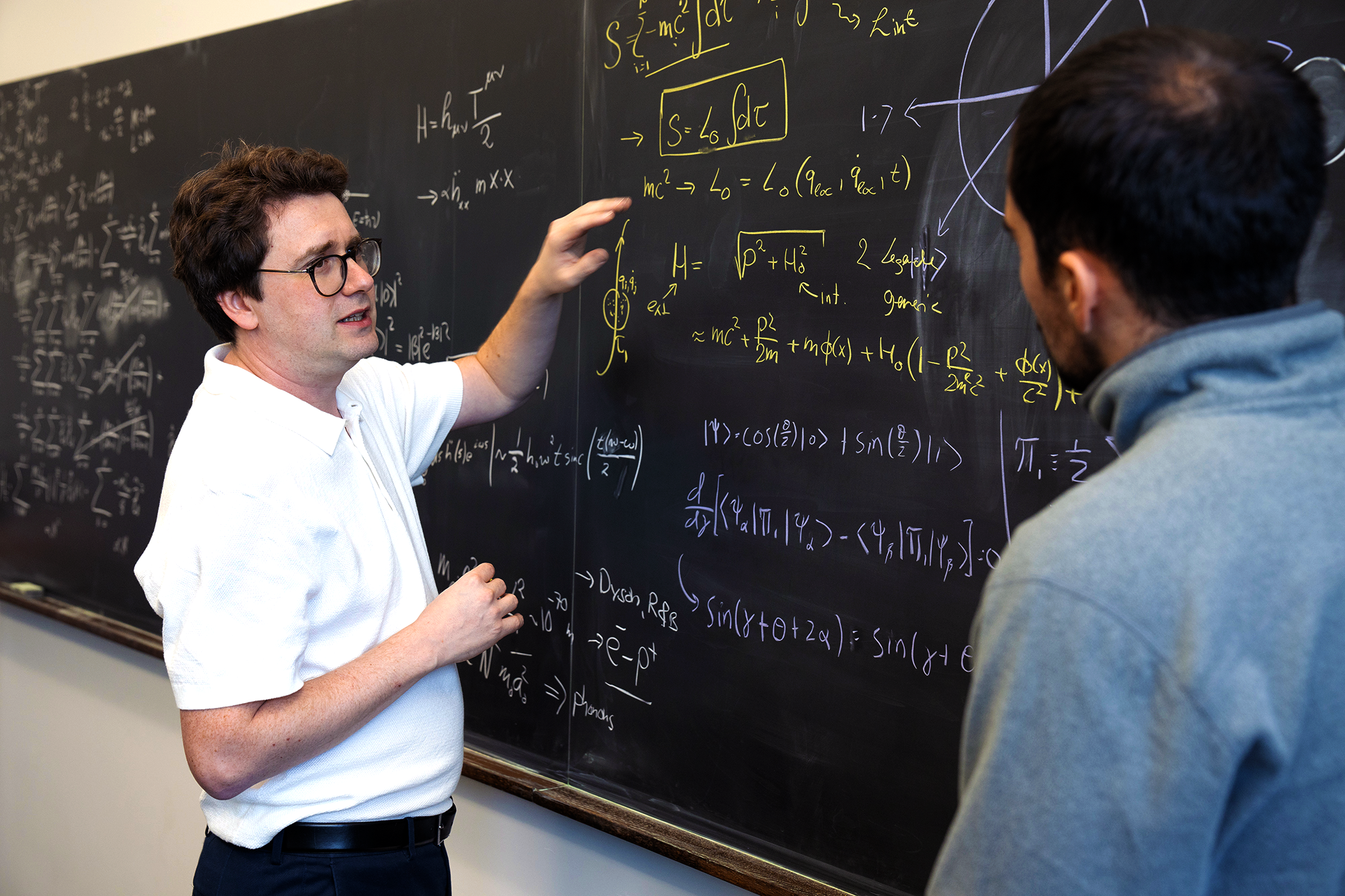 A young man in plastic-frame glasses works at a blackboard, speaking to someone who is out of focus in the foreground.