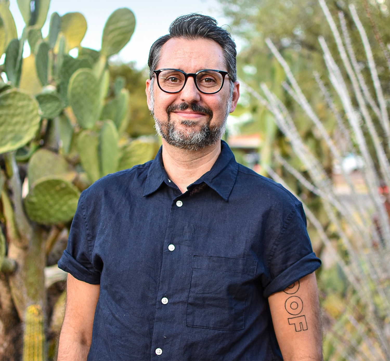 A man with glasses and a beard stands in front of a cactus.