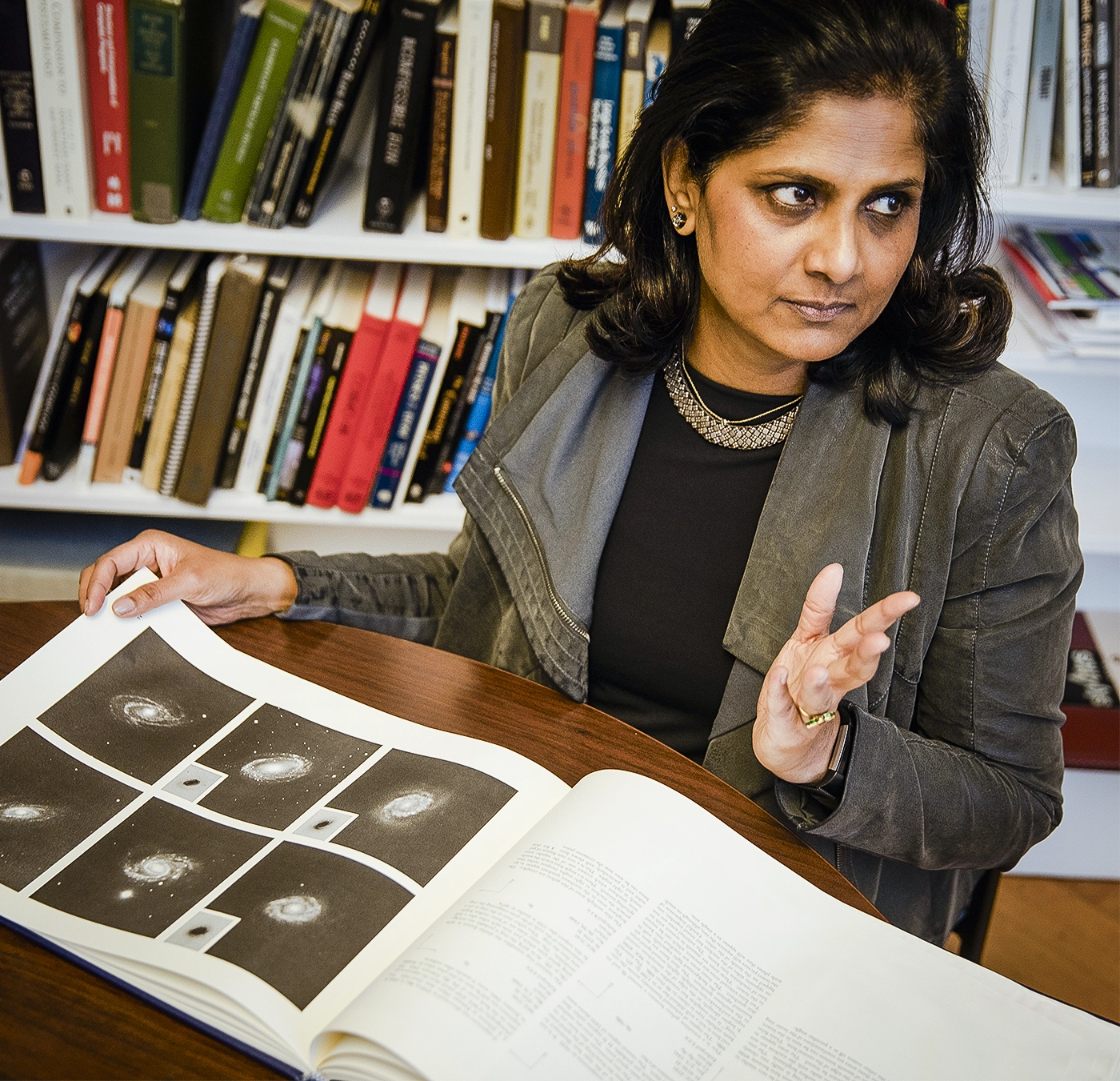 A woman gestures while flipping through a book displaying photographs of galaxies.