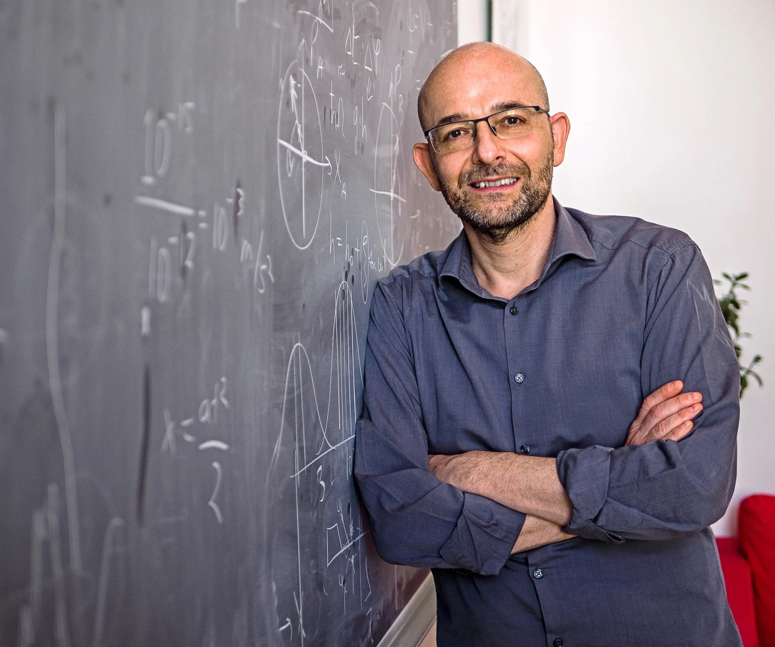 A bald, smiling man with his arms crossed leans against a blackboard.