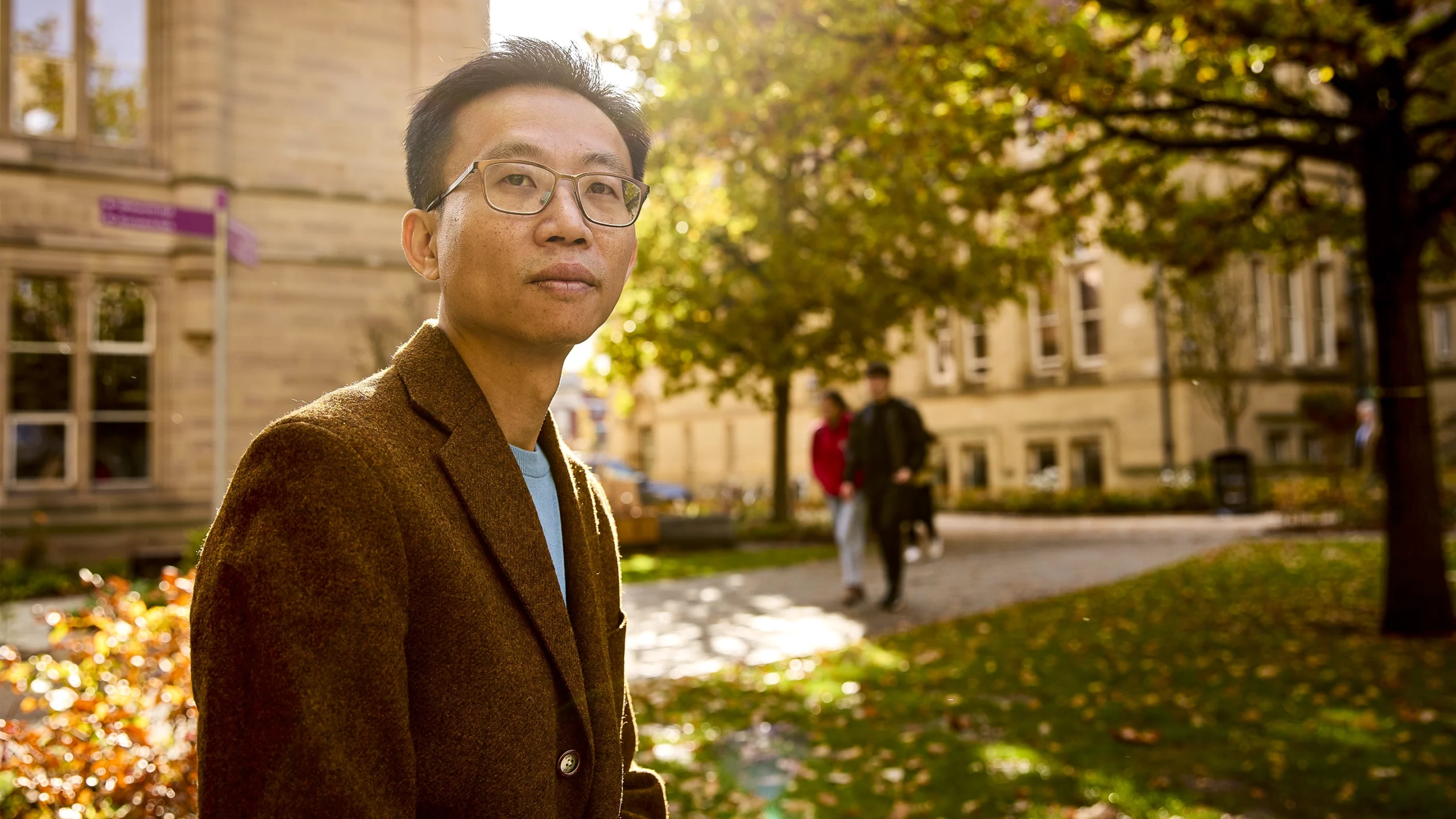 Yizhi “Patrick” Cai, a man with short black hair and broad eyeglasses, gazes into the distance on the University of Manchester campus.]