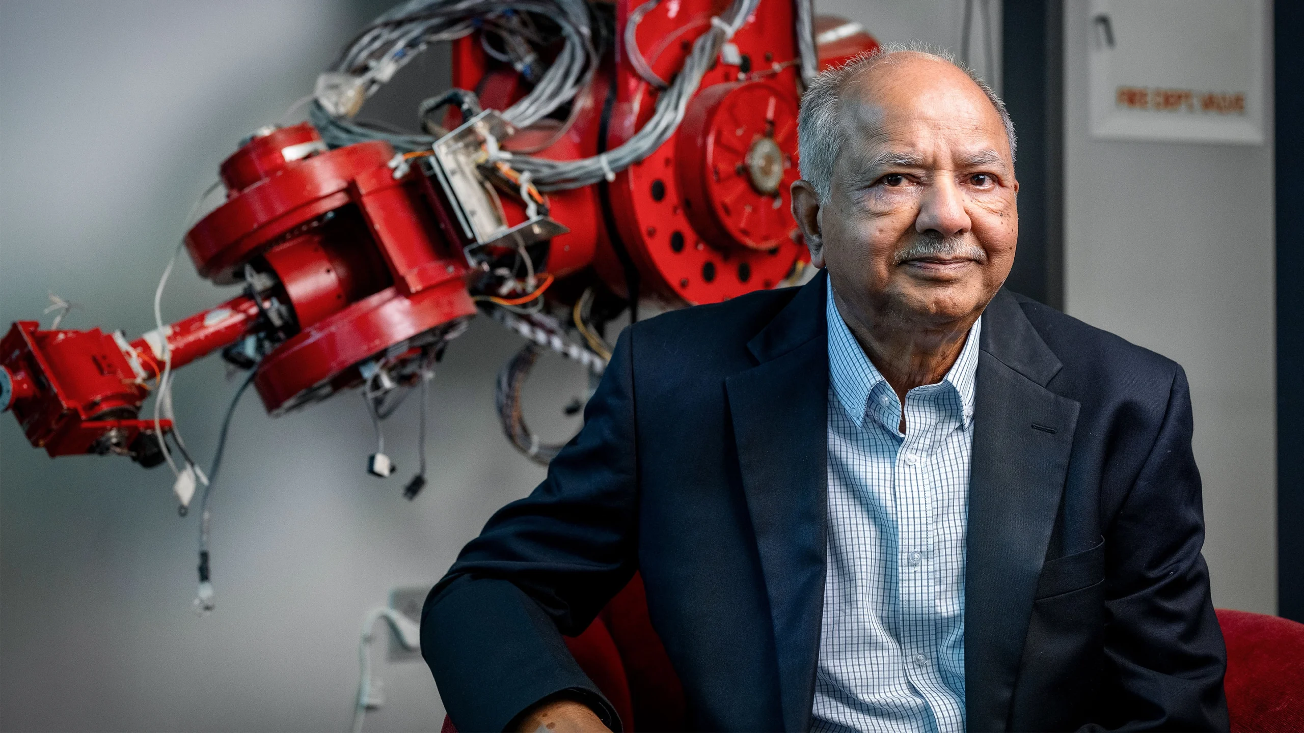 Raj Reddy in a checkered shirt and dark suit coat in front of a red metal machine.