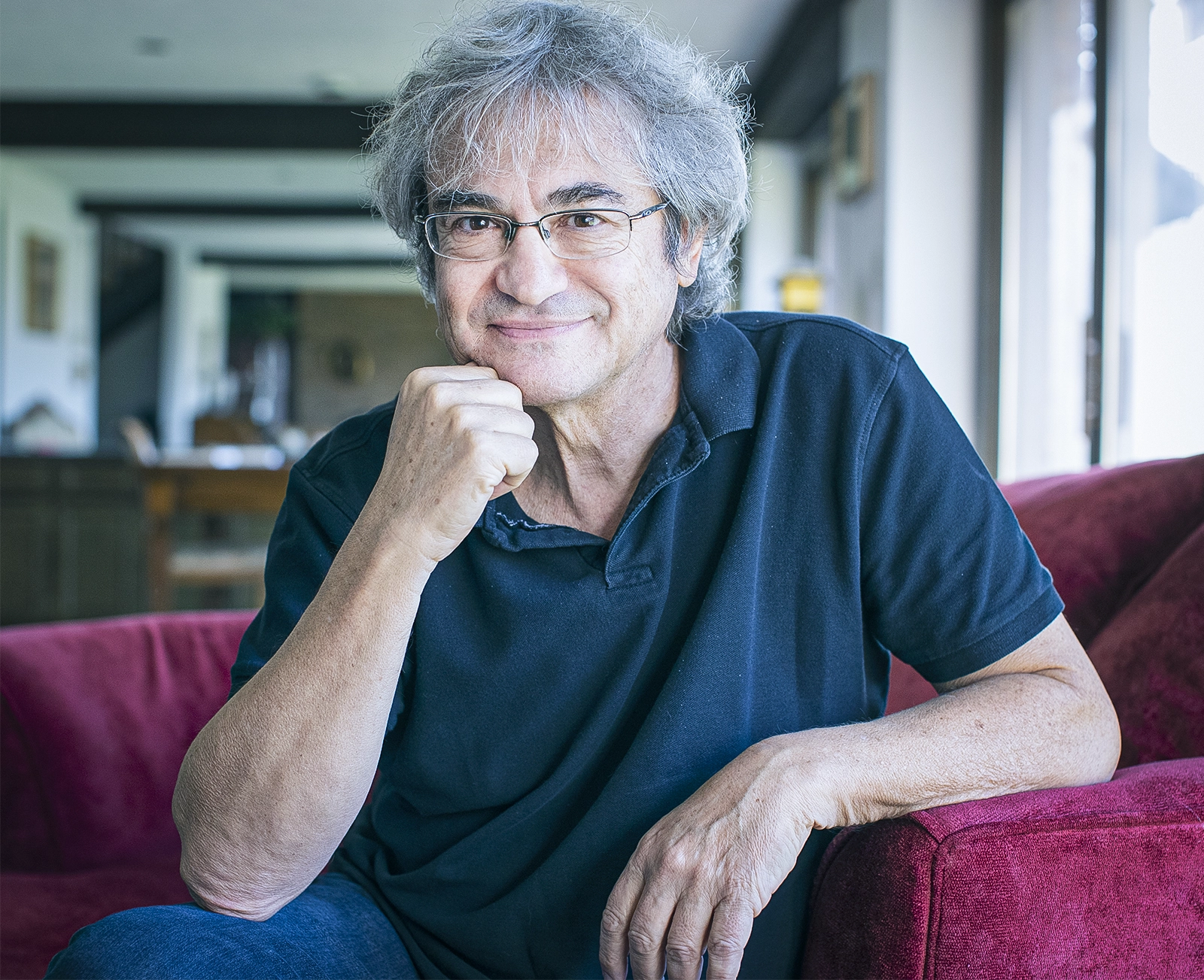 A gray-haired man in a black polo shirt sits in an armchair.