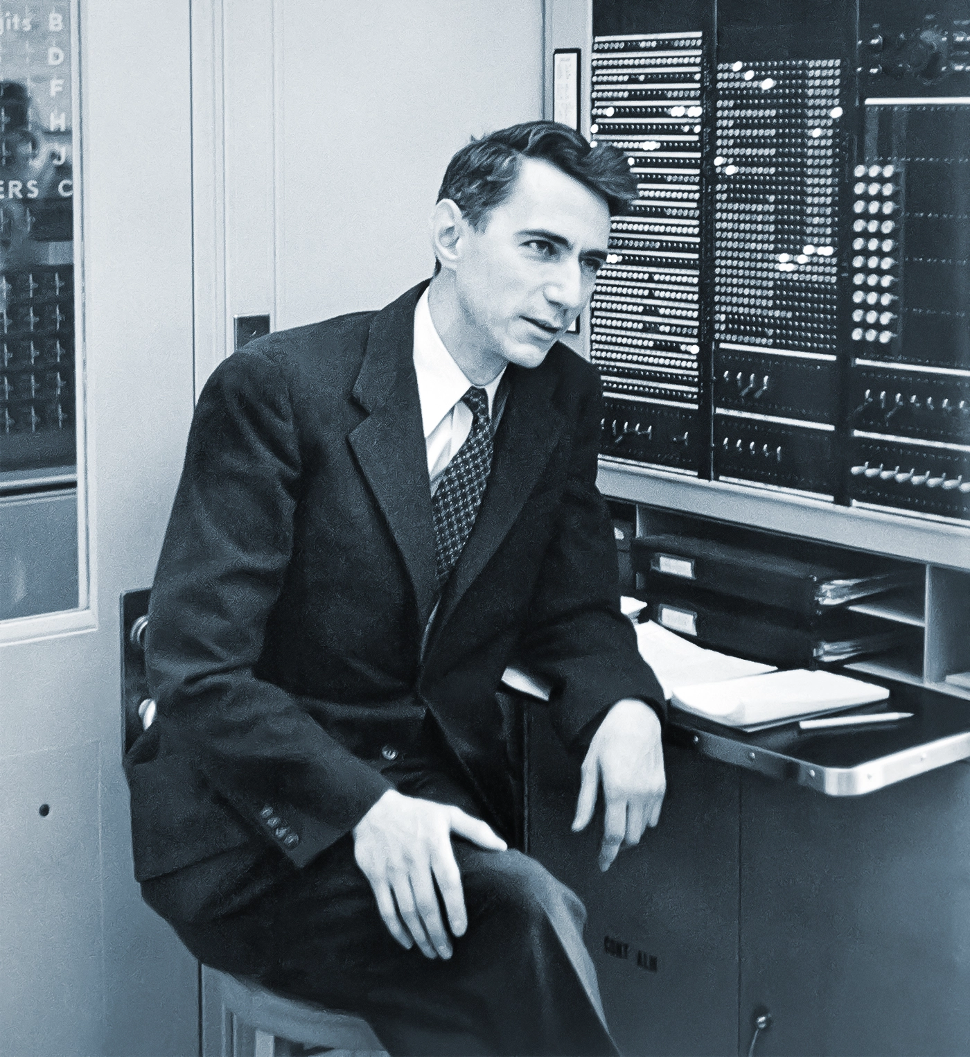 A black-and-white photo of a man in a suit sitting in front of a primitive computer.