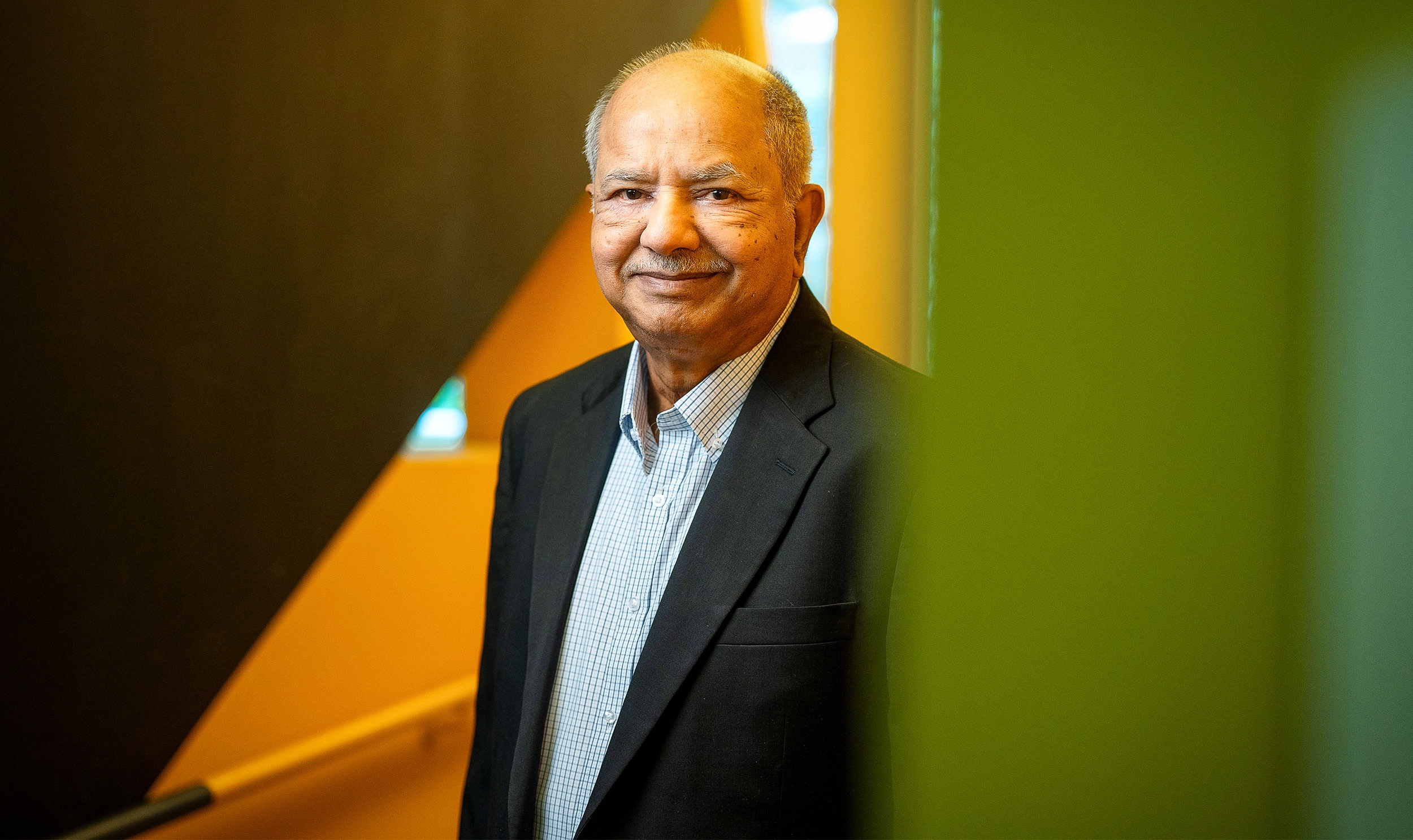 Raj Reddy in a dark suit coat stands behind a green wall and in front of an orange one.