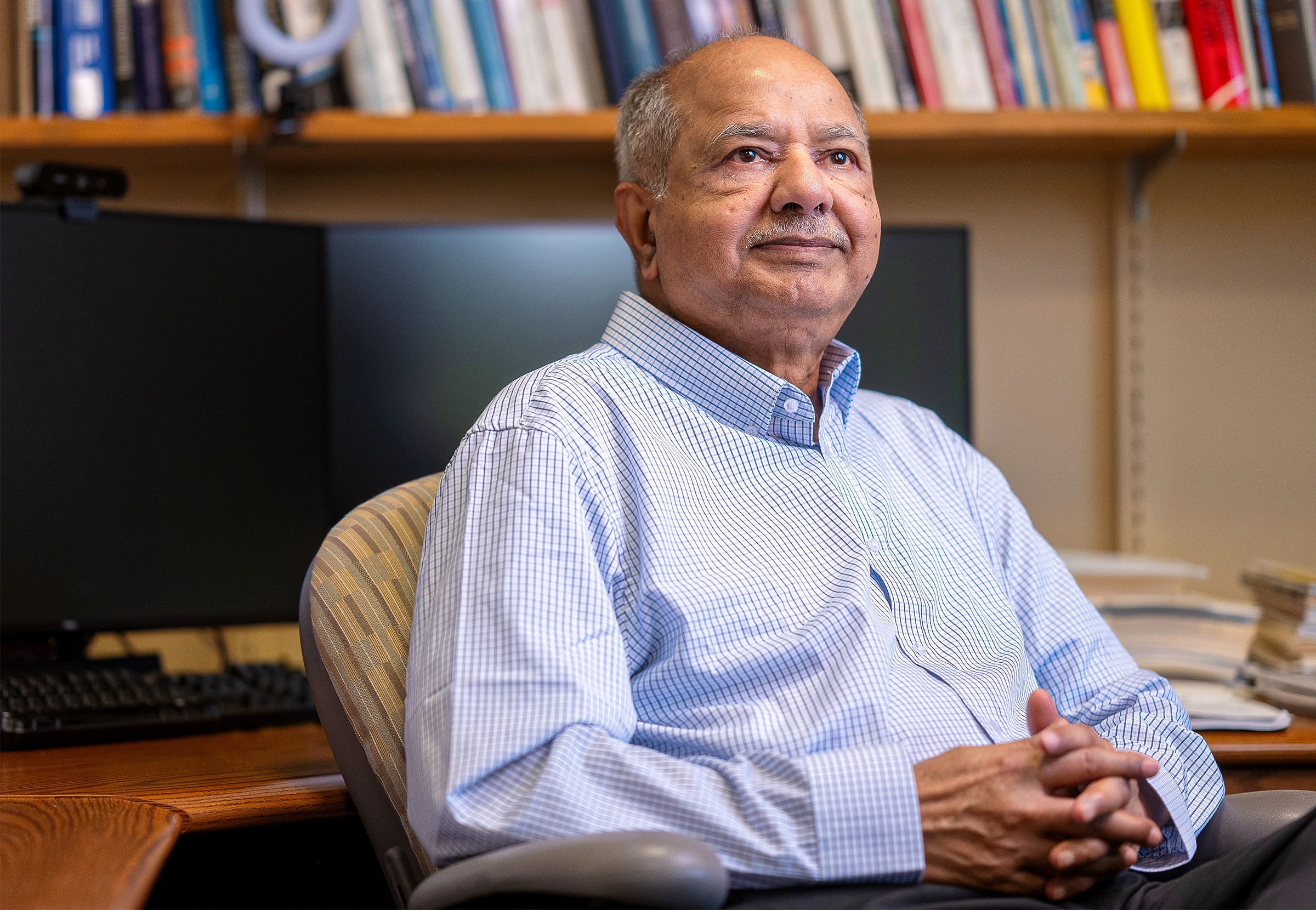 Raj Reddy in a checkered shirt sits at a desk with monitors and books behind him.