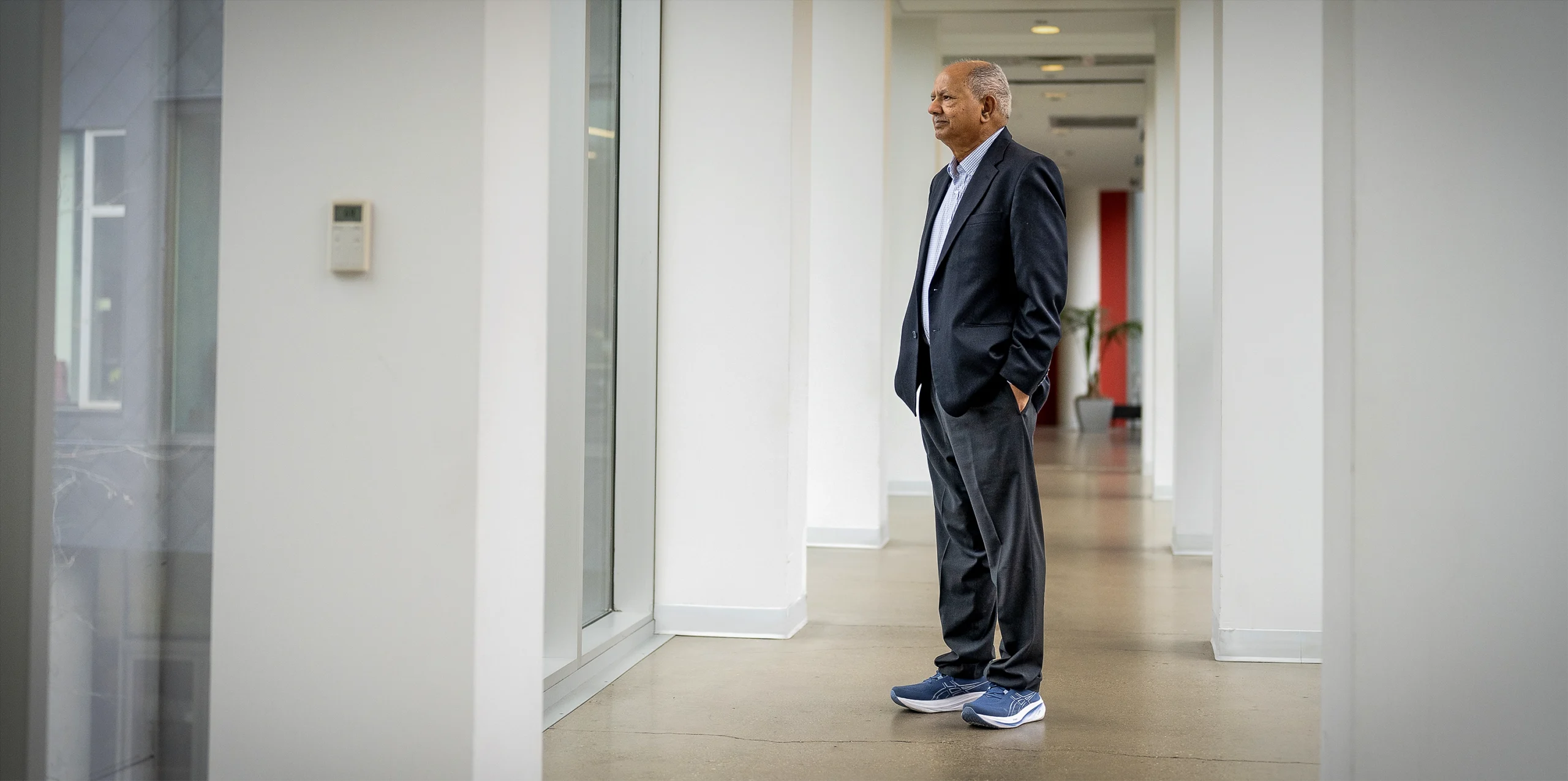 Raj Reddy stands in a suit in a white hallway, looking through a window.