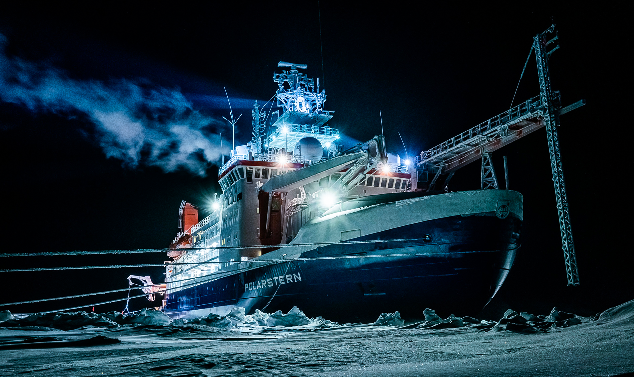 A large ship sits on an ice floe. A few bright lights from the ship illuminate the scene and reflect off the ice; otherwise the area is dark.