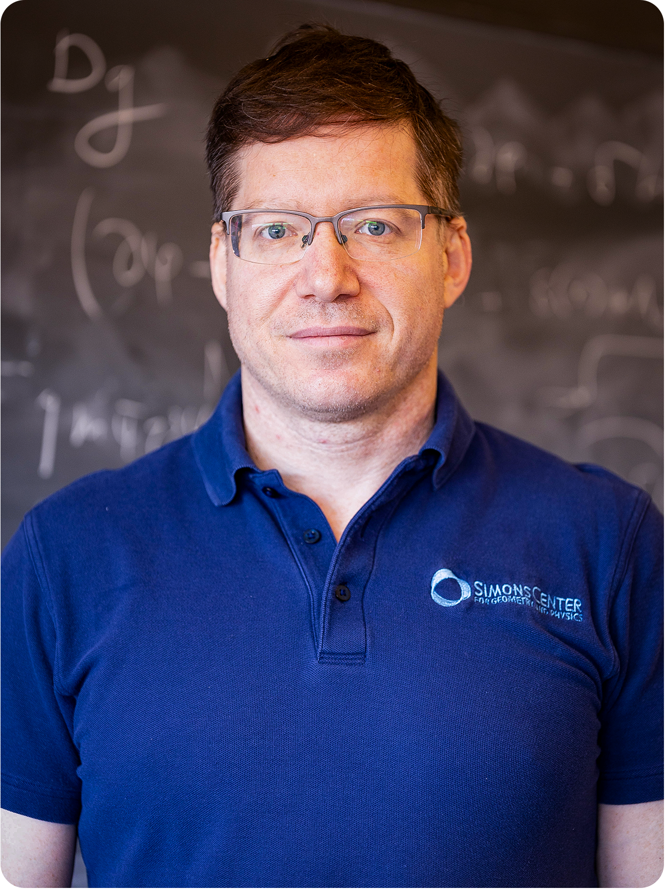 A man with glasses wearing a blue shirt and standing in front of a blackboard