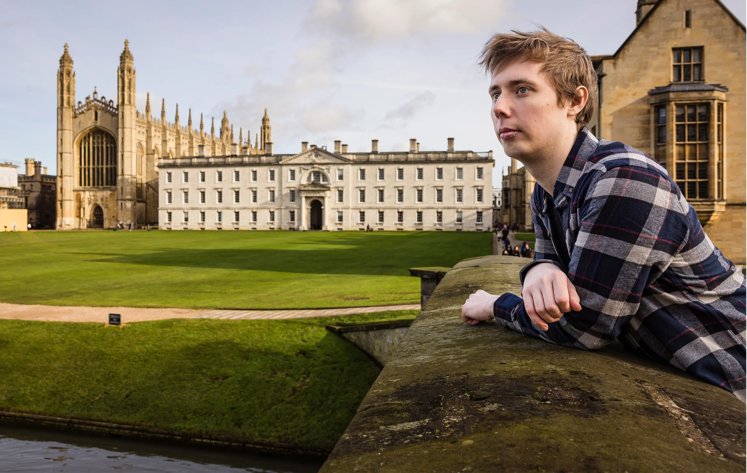 Andrew Krapivin in a blue checkered shirt leans on a bridge in front of impressive school buildings