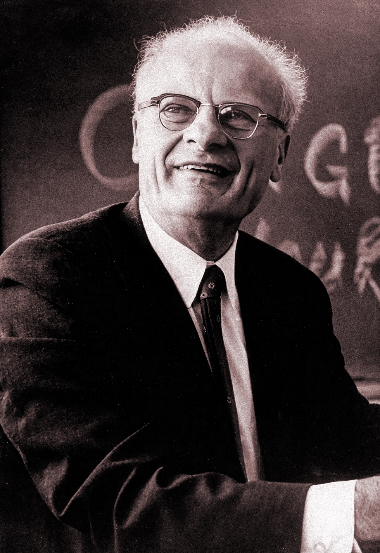 A man with wispy white hair sits in front of a blackboard.