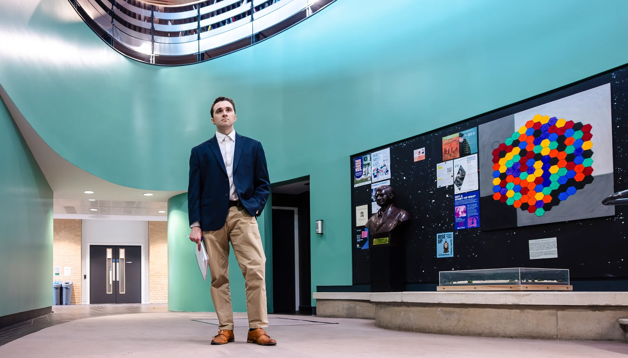 Miles Cranmer in a blue blazer and khakis stands in a large room with a science exhibit behind him