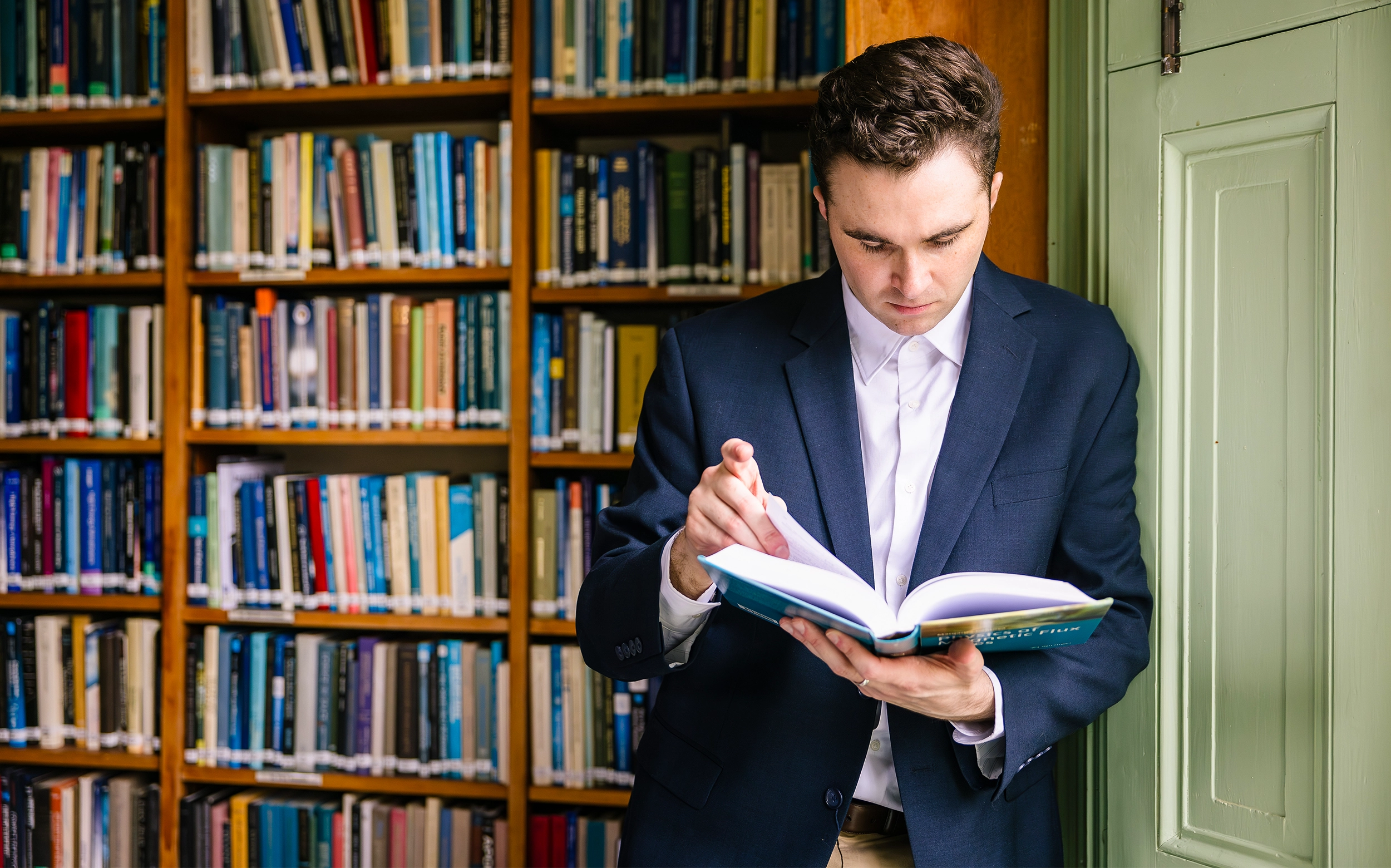 Miles Cranmer in a blue blazer looks through a book in front of a full set of bookshelves