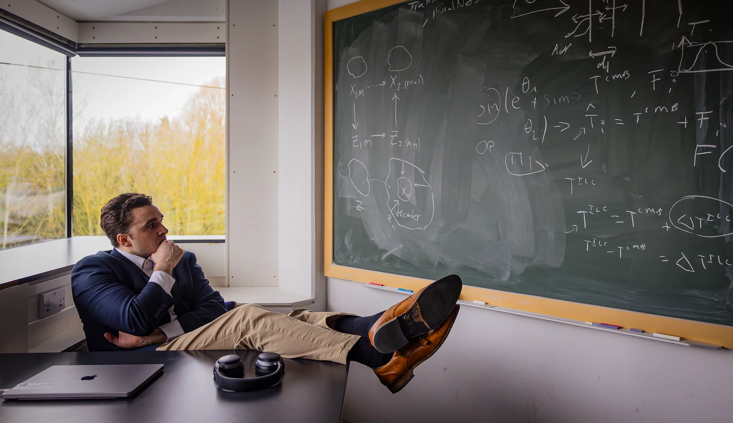 Miles Cranmer sits at a desk and looks at a chalkboard with equations