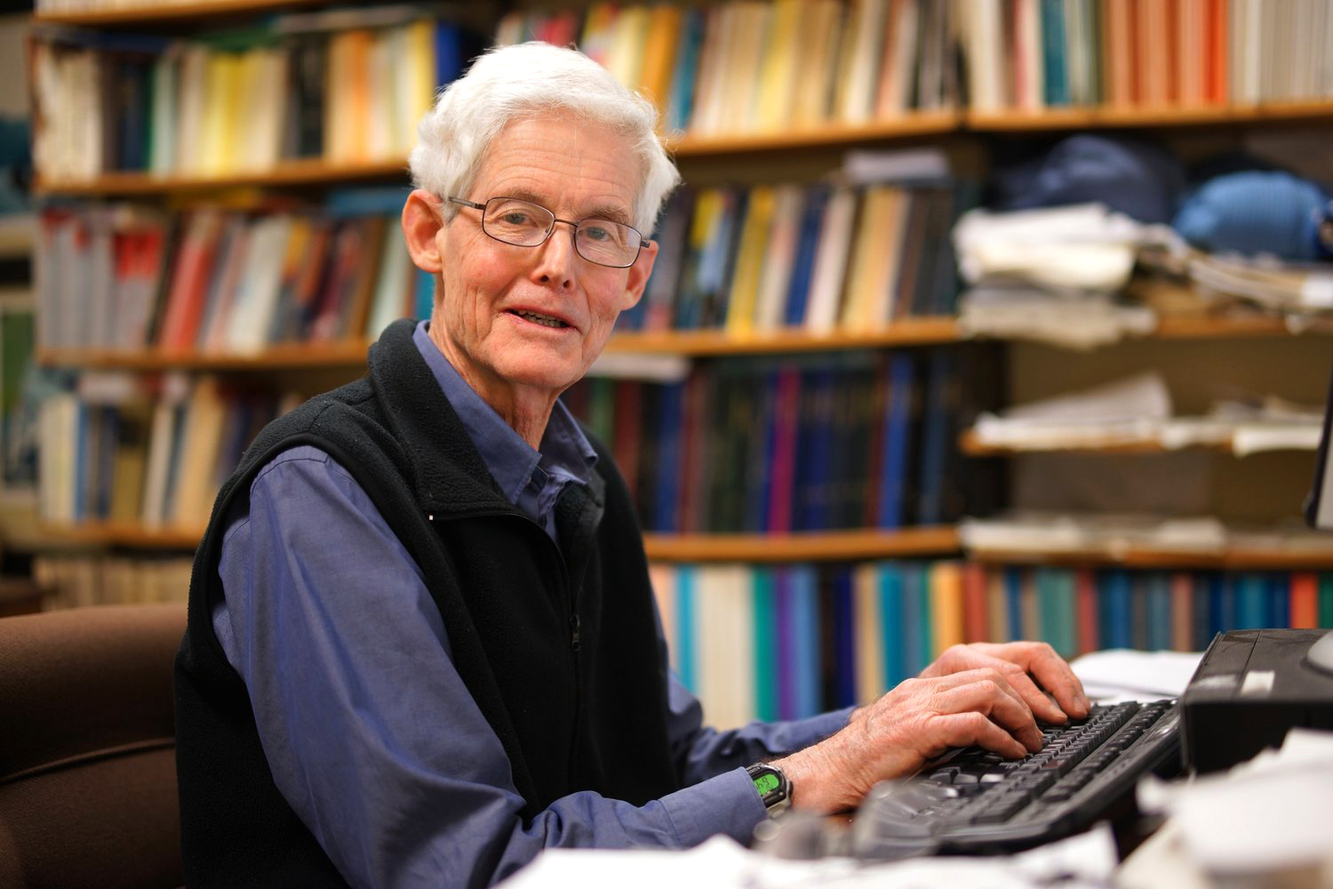 Stephen Cook in a blue shirt and black sweater types at a keyboard in front of bookshelves