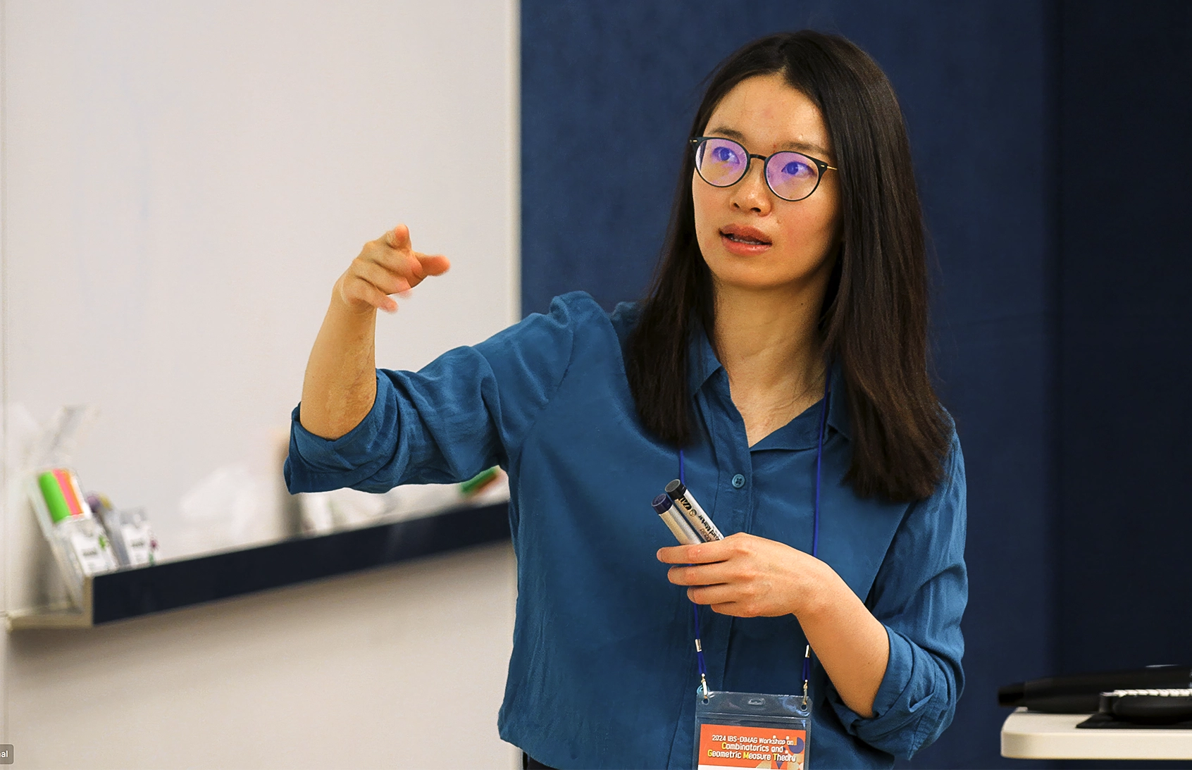 A woman in glasses lecturing in front of a white board.