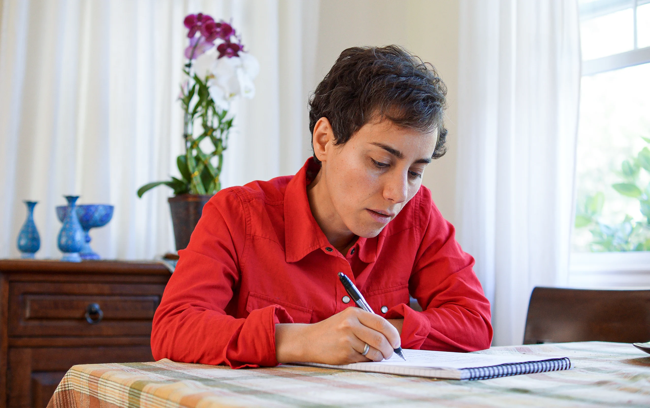 Short-haired woman in a red shirt, writing in a notebook.